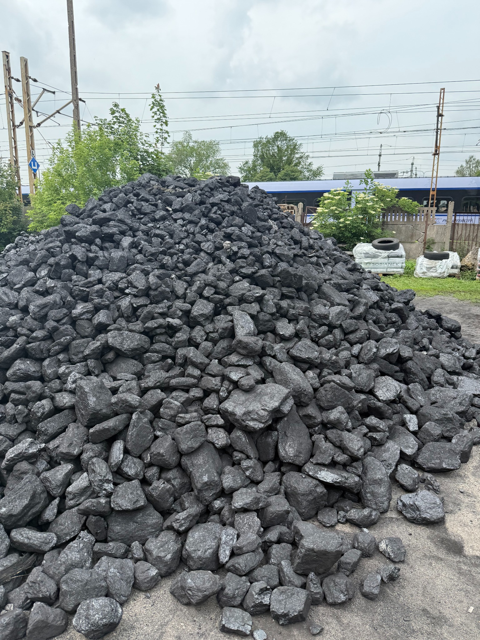 Góra czarnego węgla kamiennego na placu składowym, z widocznymi torami kolejowymi i wagonem w tle. Surowiec czeka na załadunek i transport.