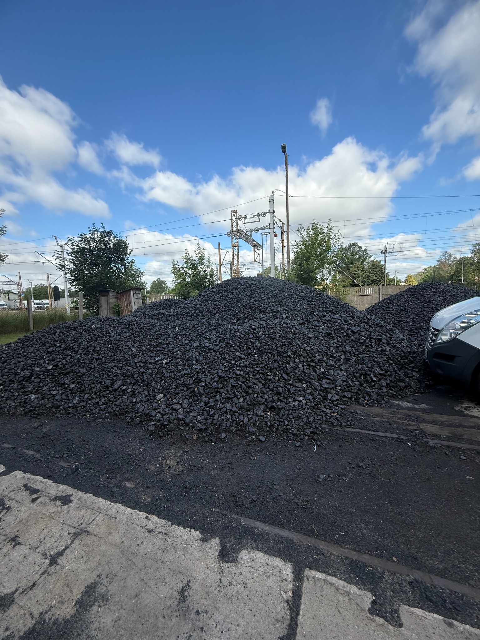 Dwa kopce czarnego opału na betonowym podłożu w słoneczny dzień, z fragmentem białego samochodu po prawej stronie. W tle słupy elektryczne i zabudowania.