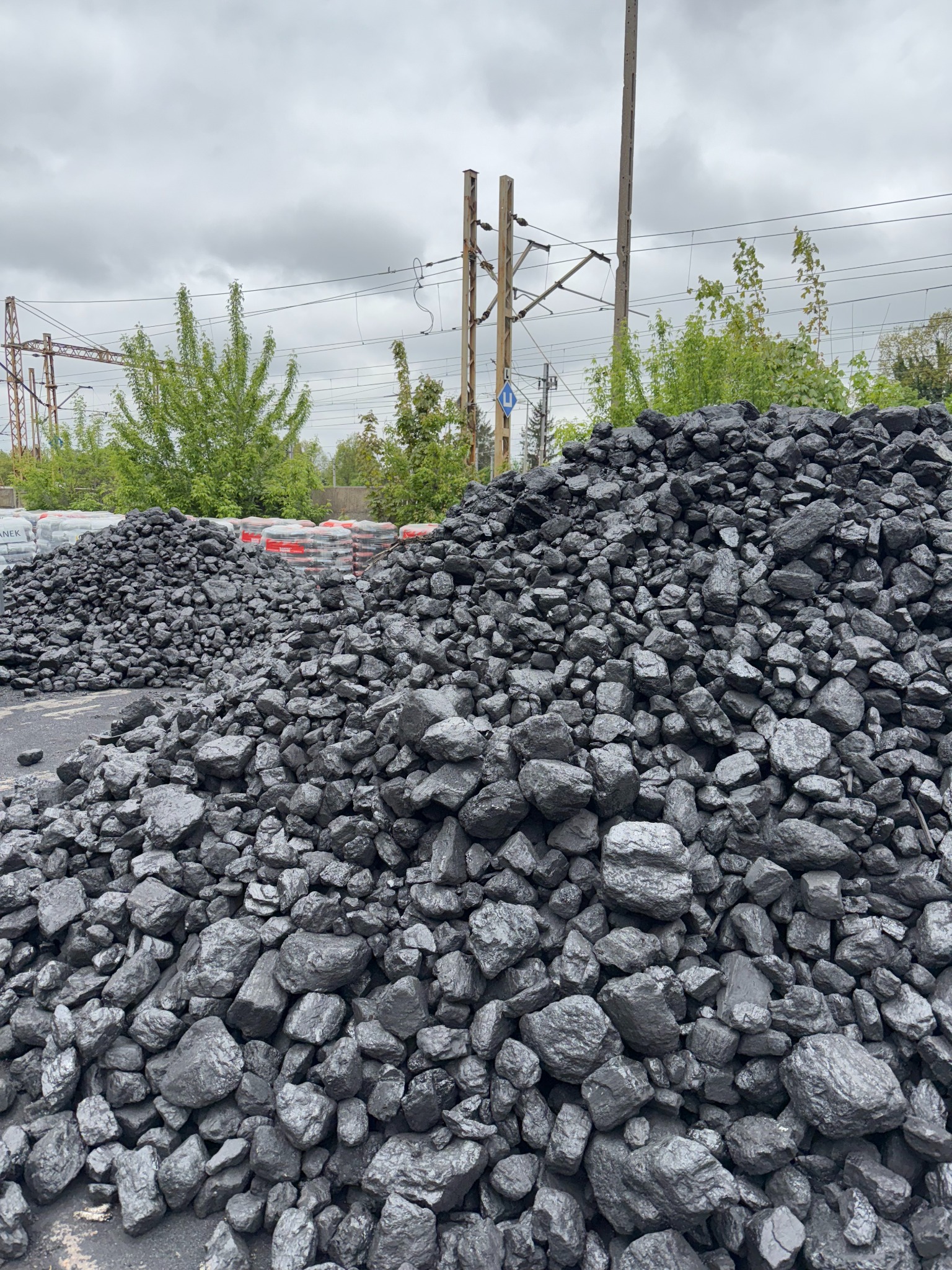 Góra czarnego węgla kamiennego na placu składowym, z widocznymi w tle workami i słupami trakcji kolejowej. Pochmurne niebo.