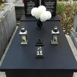 Czarny, wypolerowany nagrobek z kwiatami w wazonie i trzema lampionami. W tle inne nagrobki na cmentarzu, jesienne liście na ziemi.