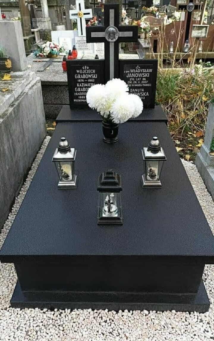 Czarny, wypolerowany nagrobek z kwiatami w wazonie i trzema lampionami. W tle inne nagrobki na cmentarzu, jesienne liście na ziemi.