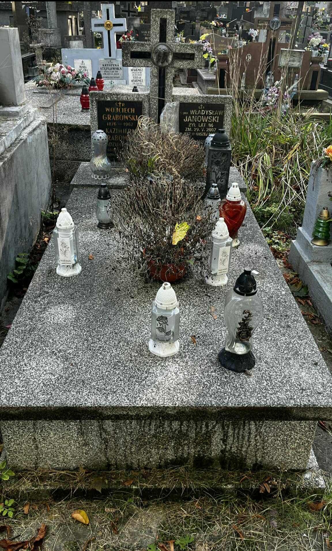 Granitowy nagrobek z wiązanką suchych gałęzi i zapalonymi zniczami na cmentarzu, widok z góry. W tle inne nagrobki i krzyże.