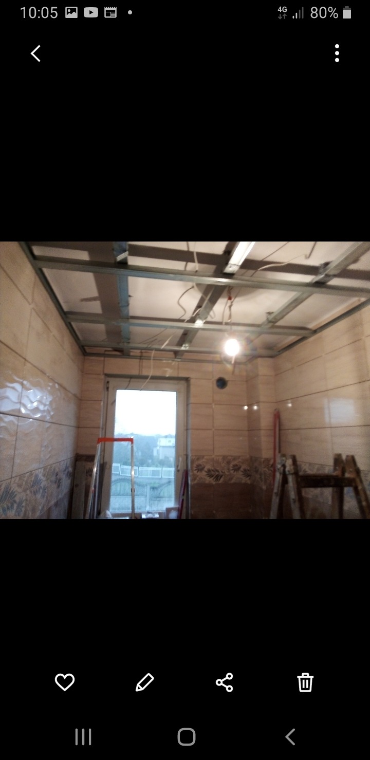 Remont łazienki w trakcie: widoczne płytki na ścianach, metalowa konstrukcja sufitu podwieszanego, okno i drabina. Przewody elektryczne zwisają z sufitu.