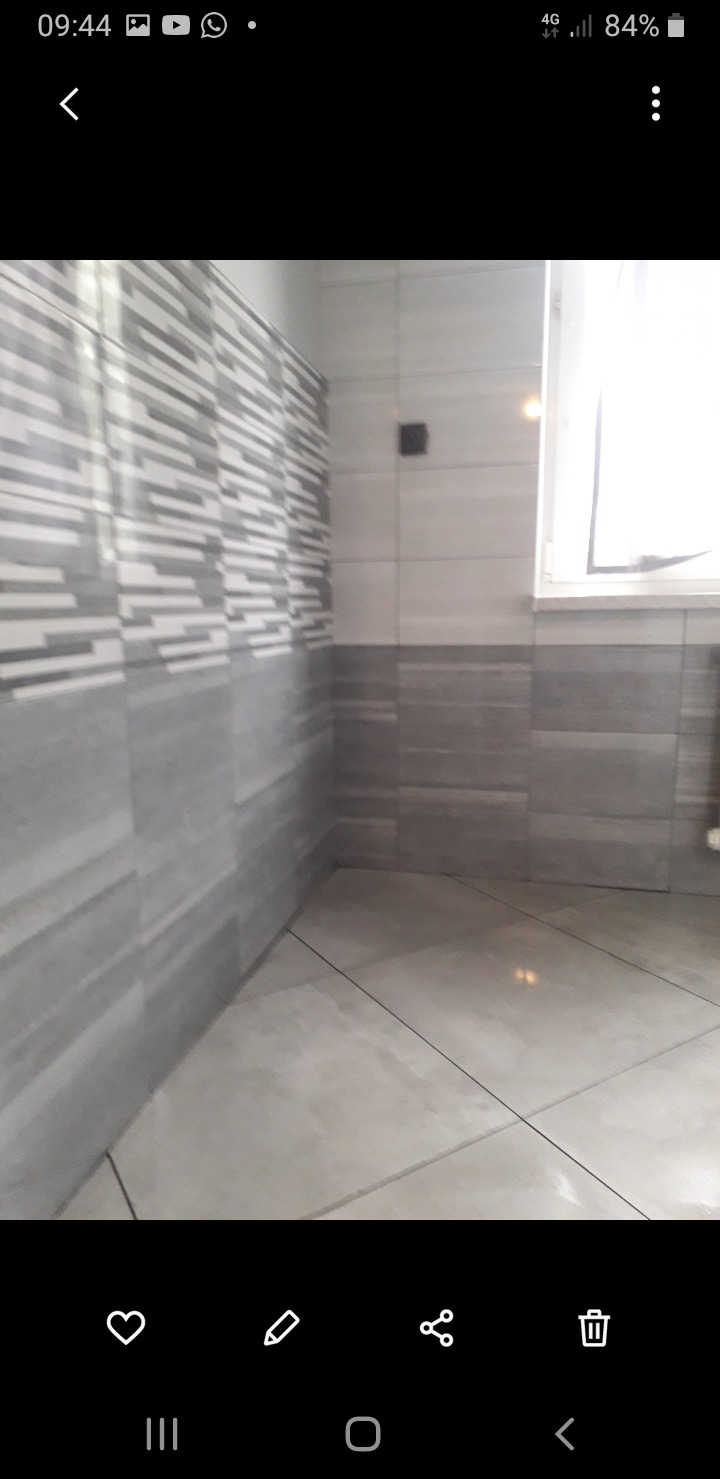 Wykończona łazienka z szarymi płytkami o zróżnicowanych wzorach na ścianach i podłodze. Widoczne fugi i okno w tle. Minimalistyczny design.