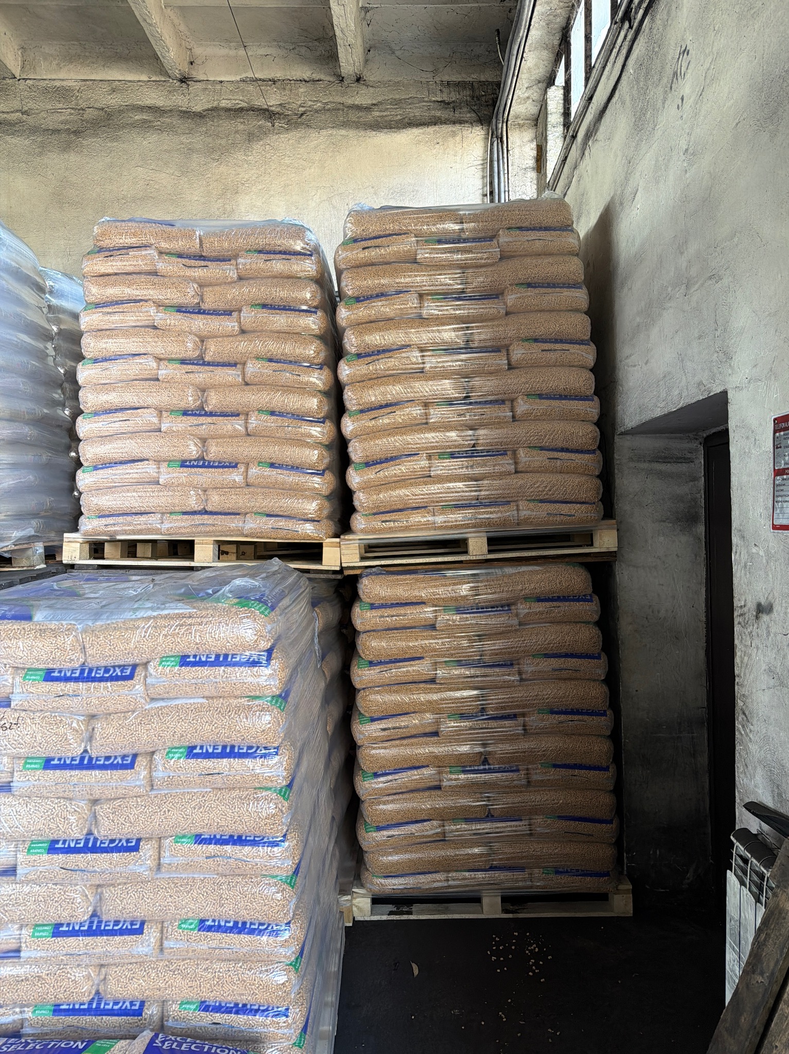 Magazyn pelletu: stosy worków na paletach, owinięte folią, z widocznym napisem 'EXCELLENT'. Surowe, betonowe ściany i podłoga. W tle drzwi i grzejnik.