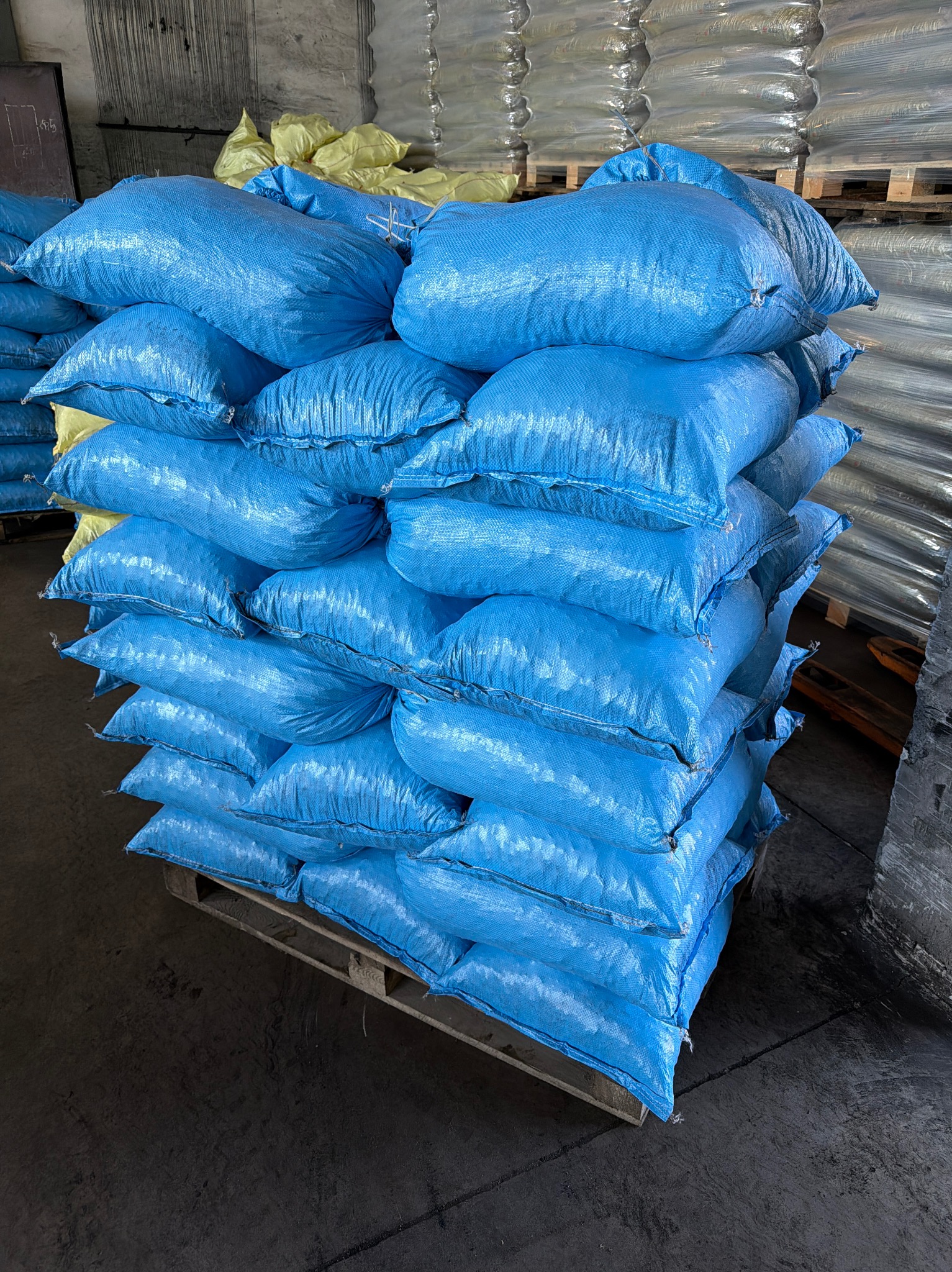 Paleta z ułożonymi workami w niebieskim kolorze. W tle widoczne inne palety z workami, niektóre w żółtym kolorze, całość w magazynie.