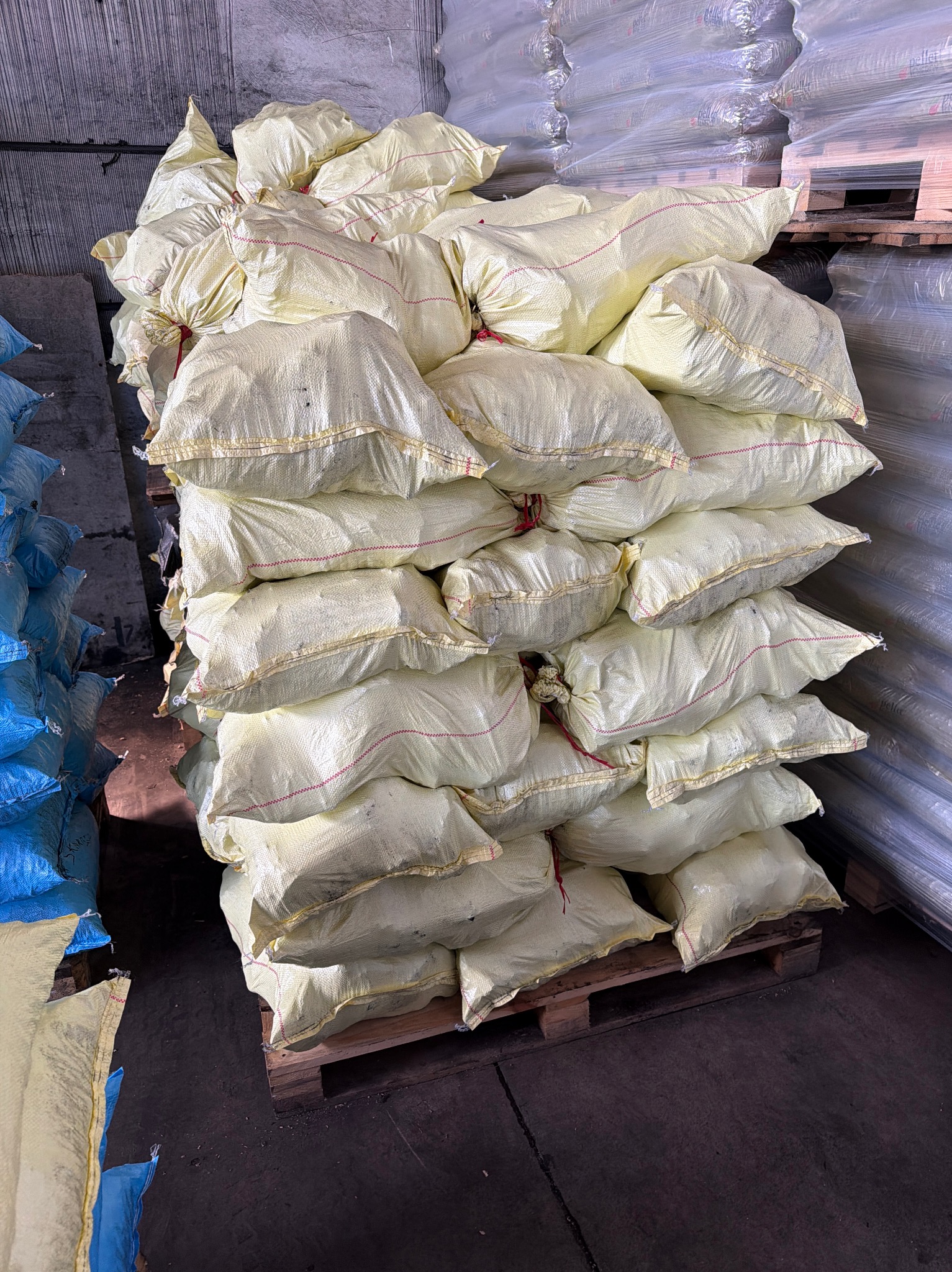 Stos worków z pelletem na palecie w magazynie. Żółte worki z czerwoną nicią, ułożone jeden na drugim. W tle widoczne inne materiały budowlane.