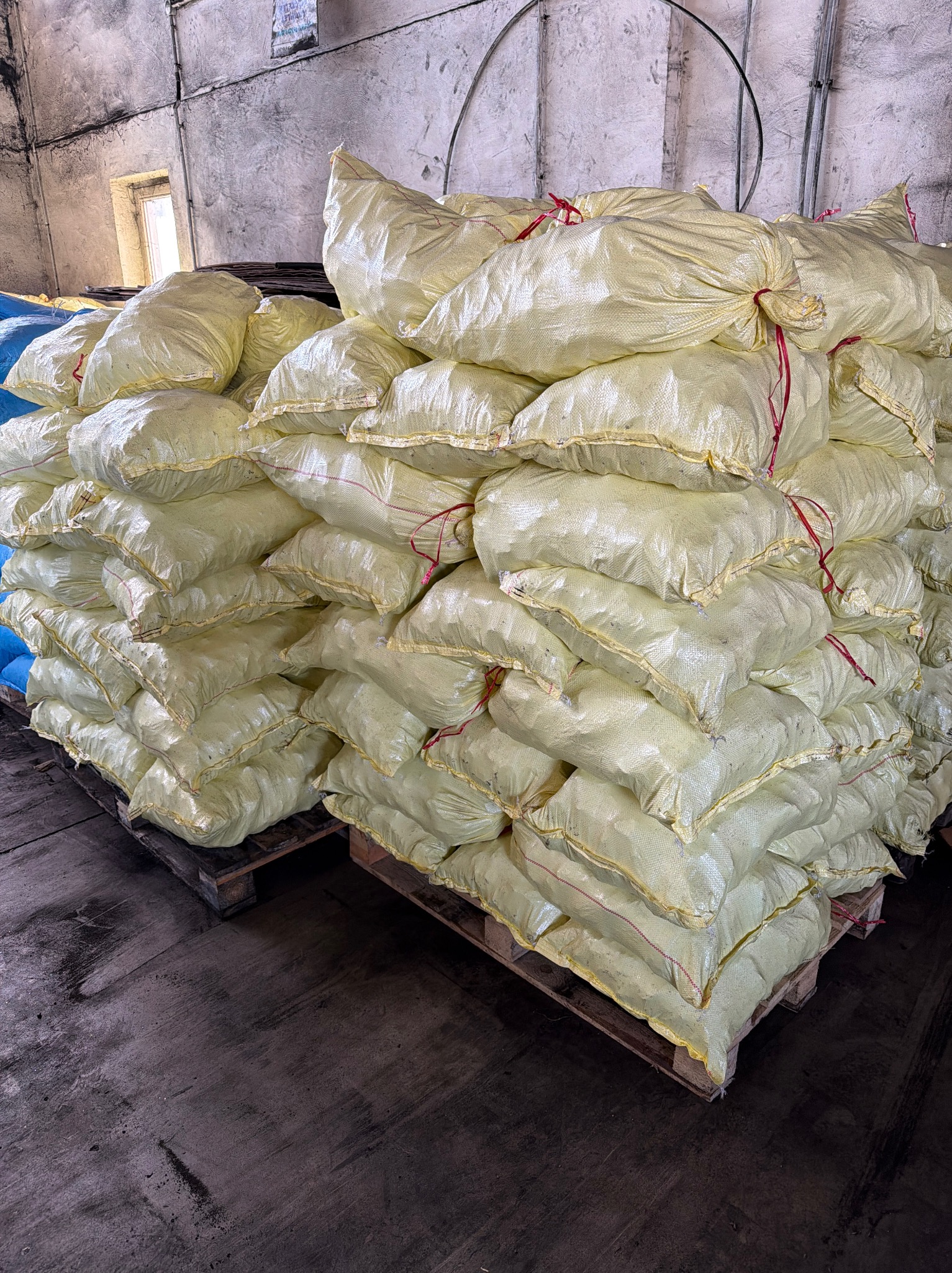 Stos worków z pelletem na palecie w magazynie. Żółte worki z czerwonym sznurkiem ułożone warstwami. Widoczne betonowe ściany i podłoga magazynu.