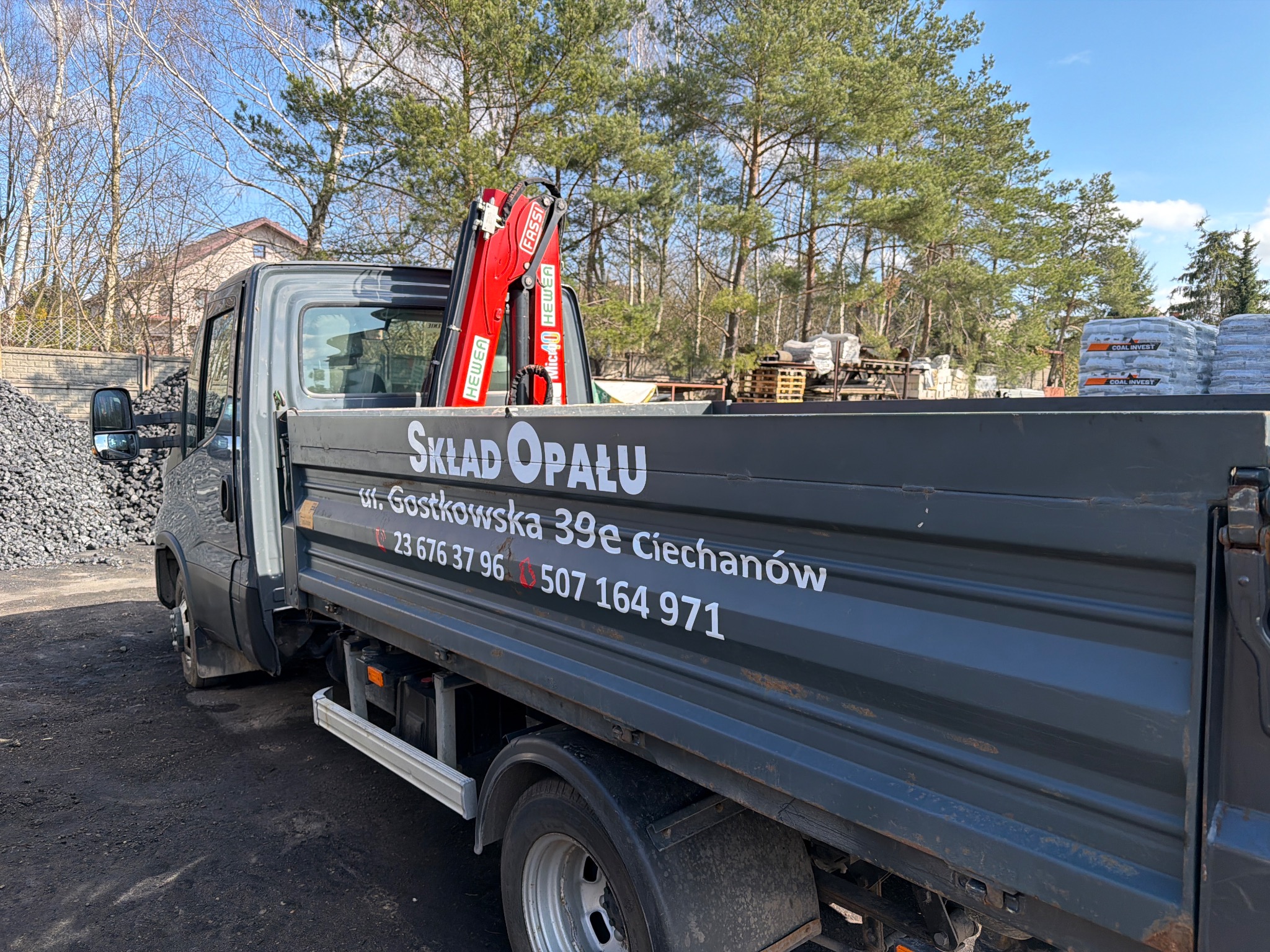 Ciężarówka z dźwigiem i napisem 'Skład Opału' na tle drzew i sterty węgla. Na pace palety i worki z napisem 'COAL INVEST'.