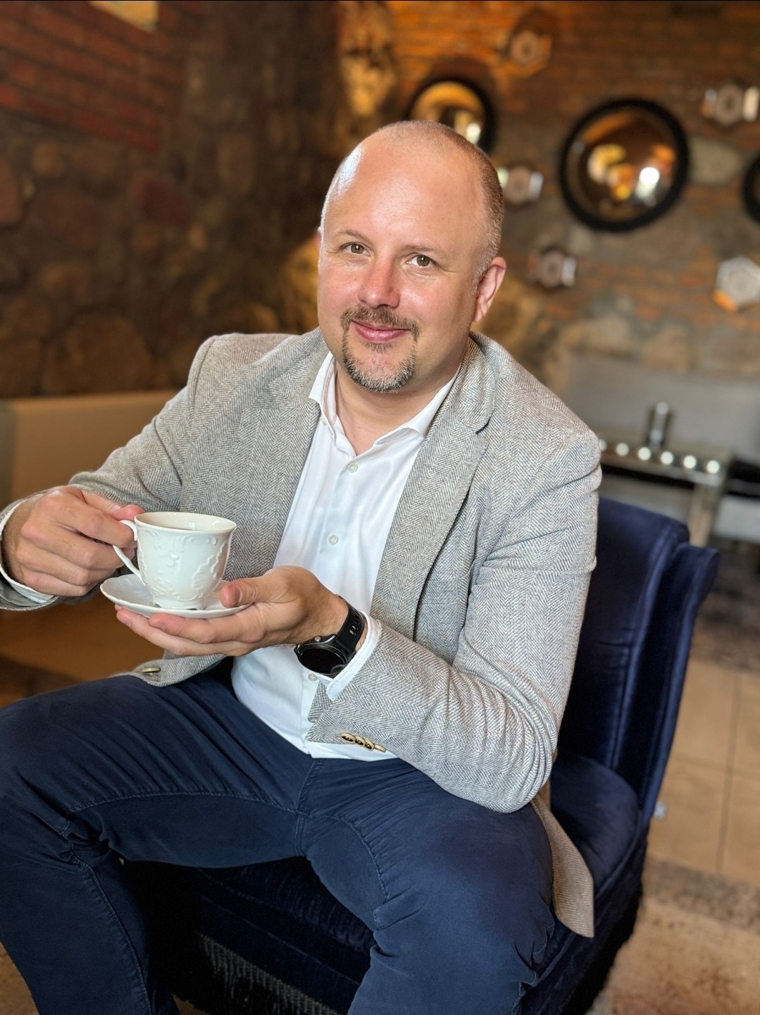 Uśmiechnięty mężczyzna w marynarce siedzi w fotelu, trzymając filiżankę. W tle dekoracyjna ściana z kamienia i luster.