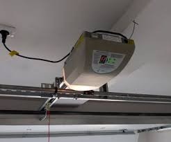 Zamontowany napęd bramy garażowej z widocznym panelem sterowania i elementami mocującymi do prowadnicy. Widoczne okablowanie i gniazdko elektryczne.