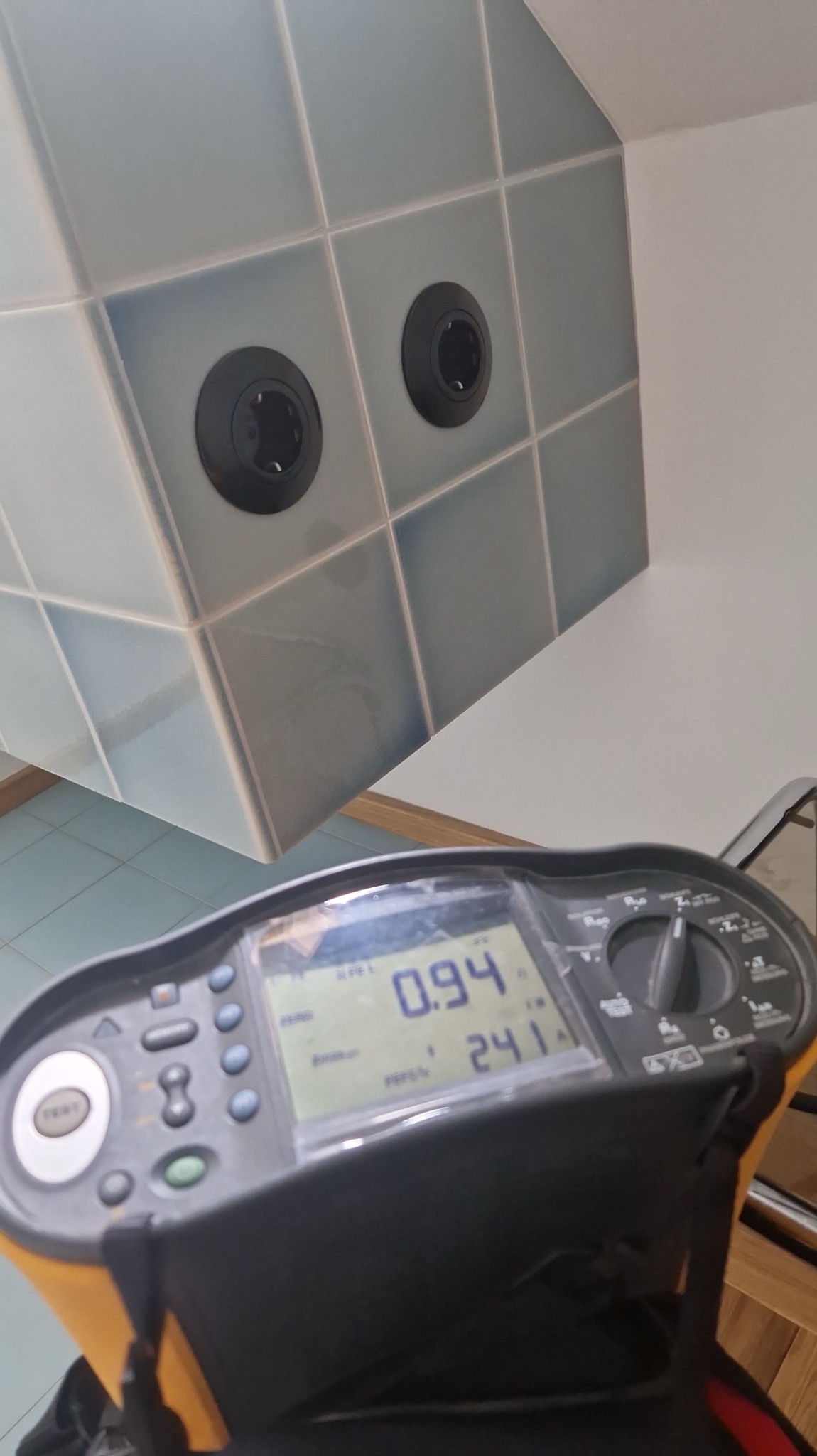 Pomiar napięcia w gniazdkach na niebieskich płytkach za pomocą miernika elektrycznego. Widoczne wartości pomiarowe na wyświetlaczu urządzenia.