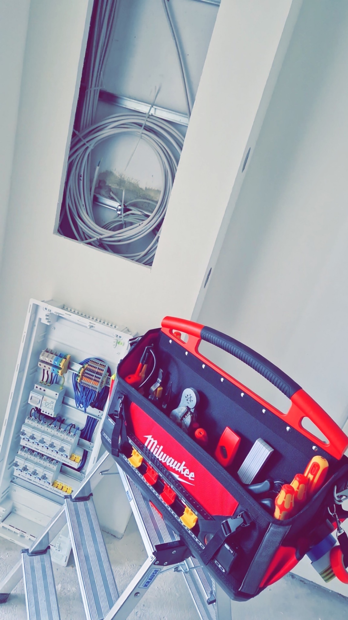 Rozdzielnica elektryczna z podłączonymi przewodami, widoczna z perspektywy drabiny. W tle otwór w suficie z kablami. Torba narzędziowa Milwaukee na pierwszym planie.