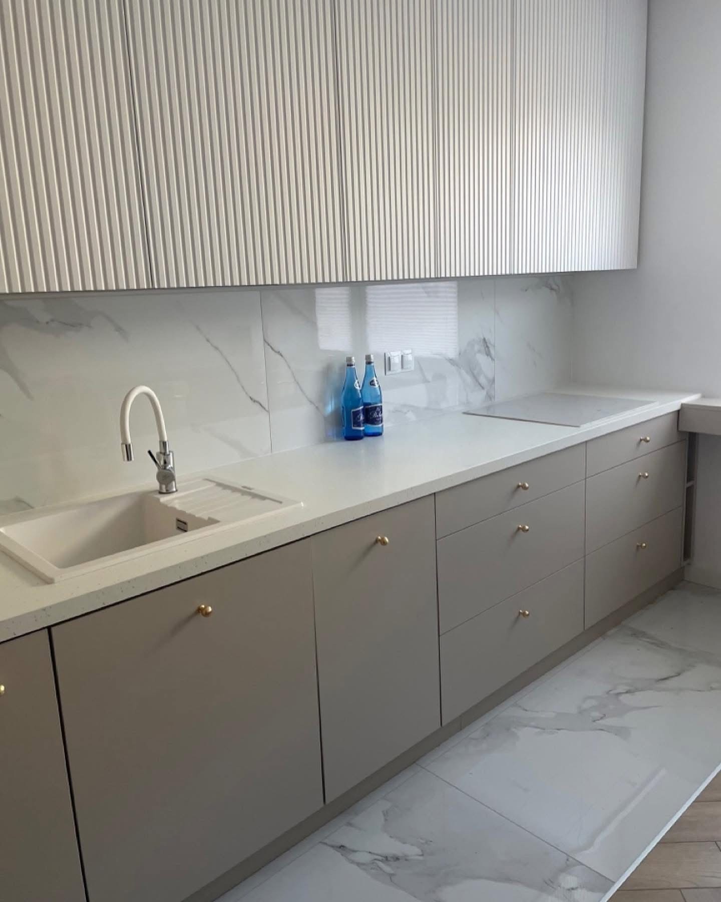 Beżowa kuchnia na wymiar z frezowanymi górnymi szafkami i białym blatem. Złote gałki, marmurowe płytki na ścianie i podłodze. Dwie niebieskie butelki na blacie.