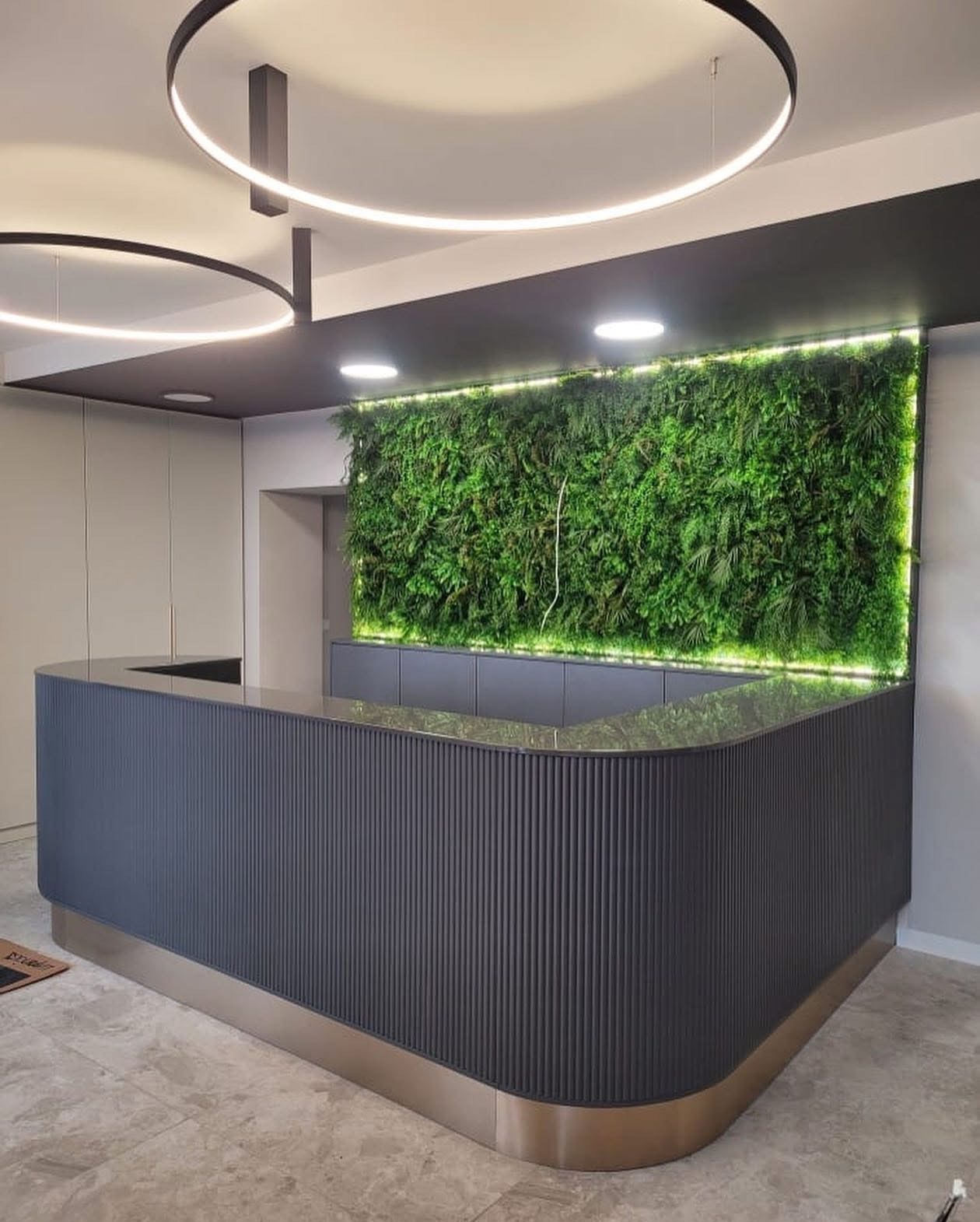 Recepcja z szarą, ryflowaną zabudową i blatem, podświetlona zielona ściana z roślin, okrągłe lampy sufitowe. Nowoczesny design wnętrza biurowego.