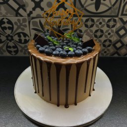 Ciacho Ciach - Pracownia Wypieków - Tort urodzinowy z borówkami, kawałkami czekolady i napisem 'Happy Birthday' na tle mozaiki. Kremowy tort z polewą czekoladową na białym talerzu.