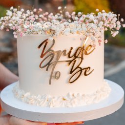 Ciacho Ciach - Pracownia Wypieków - Delikatny tort ślubny z napisem 'Bride to Be' w złotym kolorze, udekorowany drobnymi, białymi kwiatami gipsówki. Tort trzymany w dłoniach na tle zieleni.