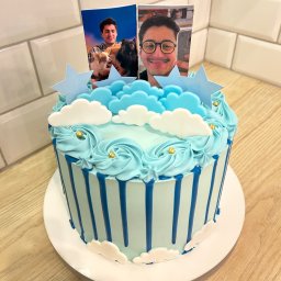 Ciacho Ciach - Pracownia Wypieków - Niebieski tort z dekoracją chmur, gwiazd i zdjęciami osób, na drewnianym blacie i tle z białych płytek.