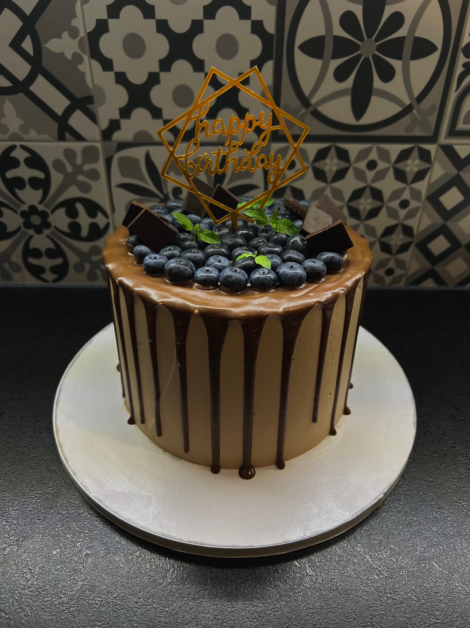 Tort urodzinowy z borówkami, kawałkami czekolady i napisem 'Happy Birthday' na tle mozaiki. Kremowy tort z polewą czekoladową na białym talerzu.