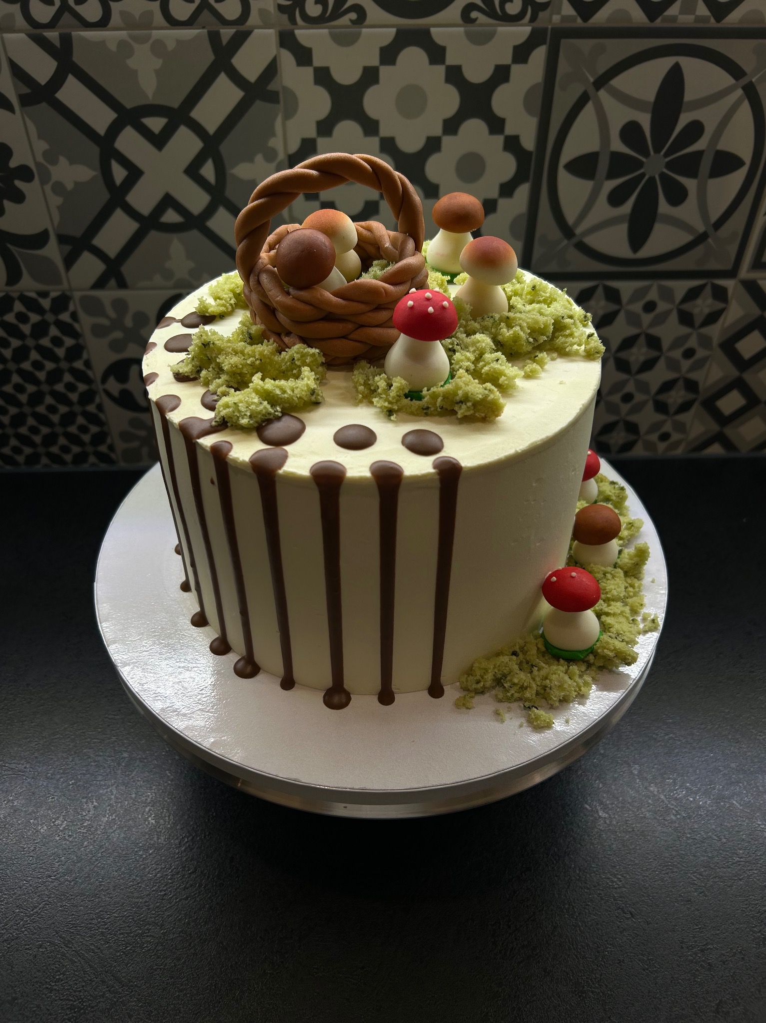 Tort z motywem grzybowym: biały krem, czekoladowe polewy, dekoracja z koszyka i grzybów z masy cukrowej na tle wzorzystych płytek.