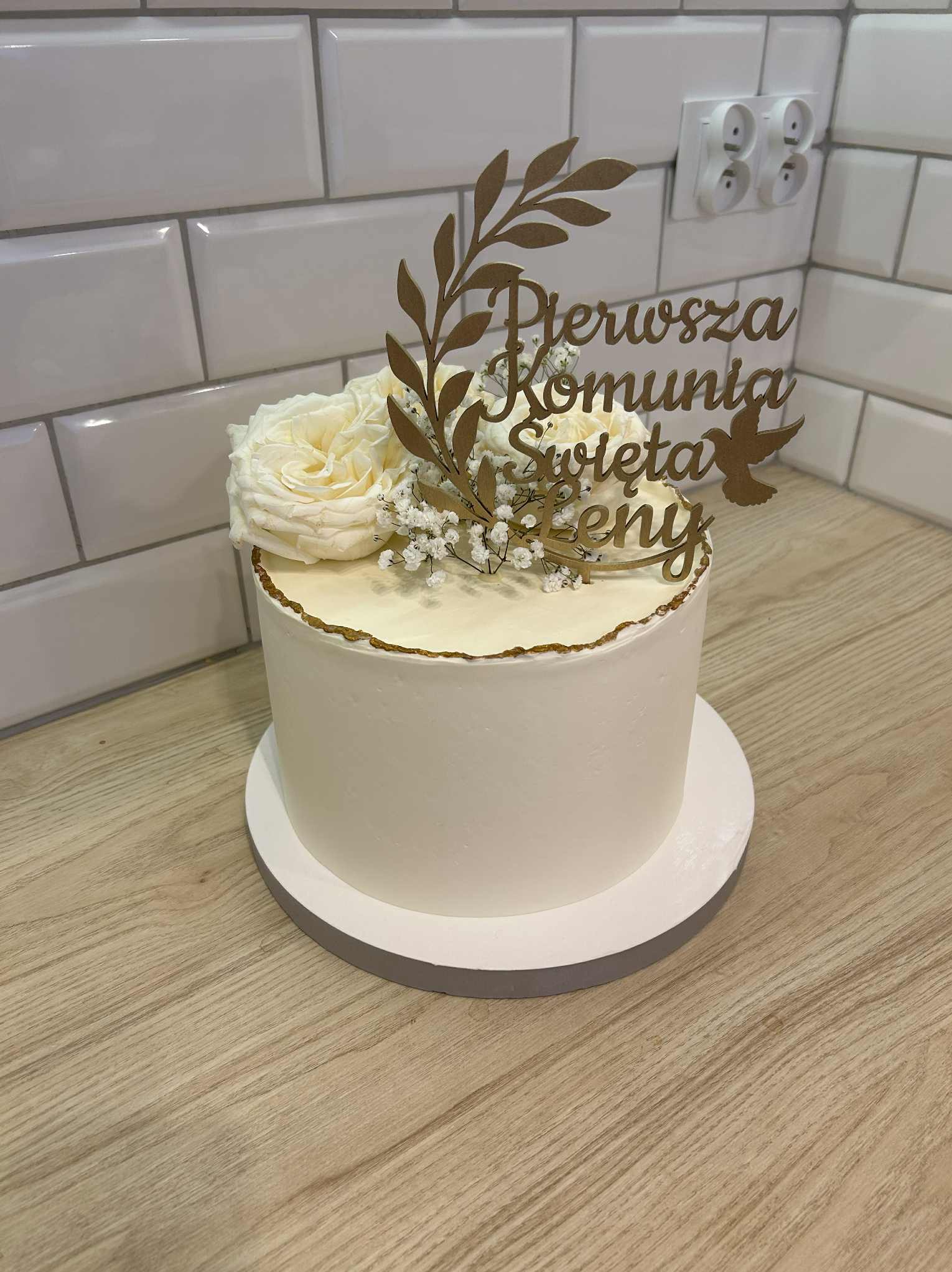 Tort komunijny z białym kremem, złotym napisem i dekoracją z białych róż i gipsówki. Minimalistyczny design na tle białych płytek.