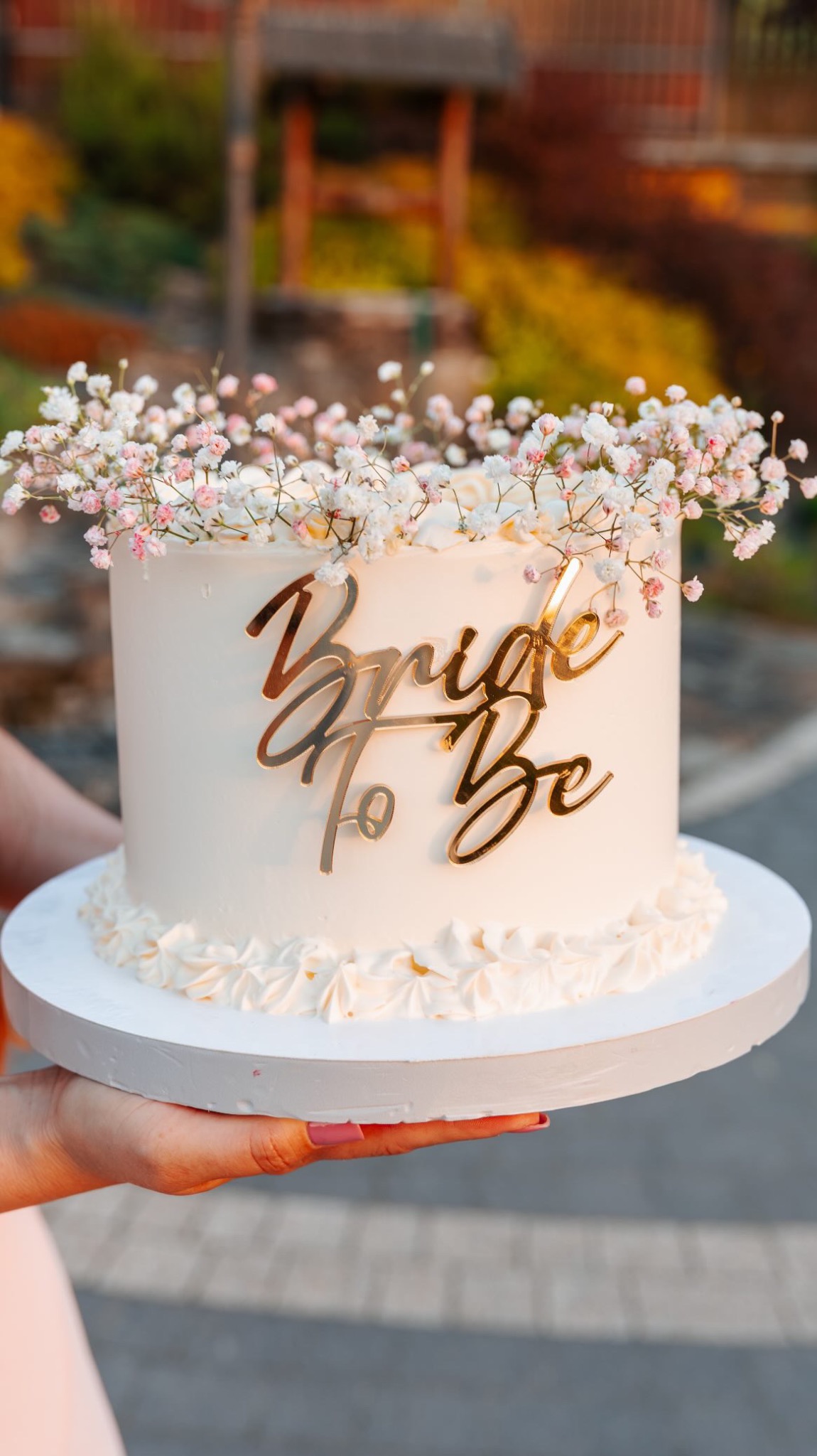 Delikatny tort ślubny z napisem 'Bride to Be' w złotym kolorze, udekorowany drobnymi, białymi kwiatami gipsówki. Tort trzymany w dłoniach na tle zieleni.