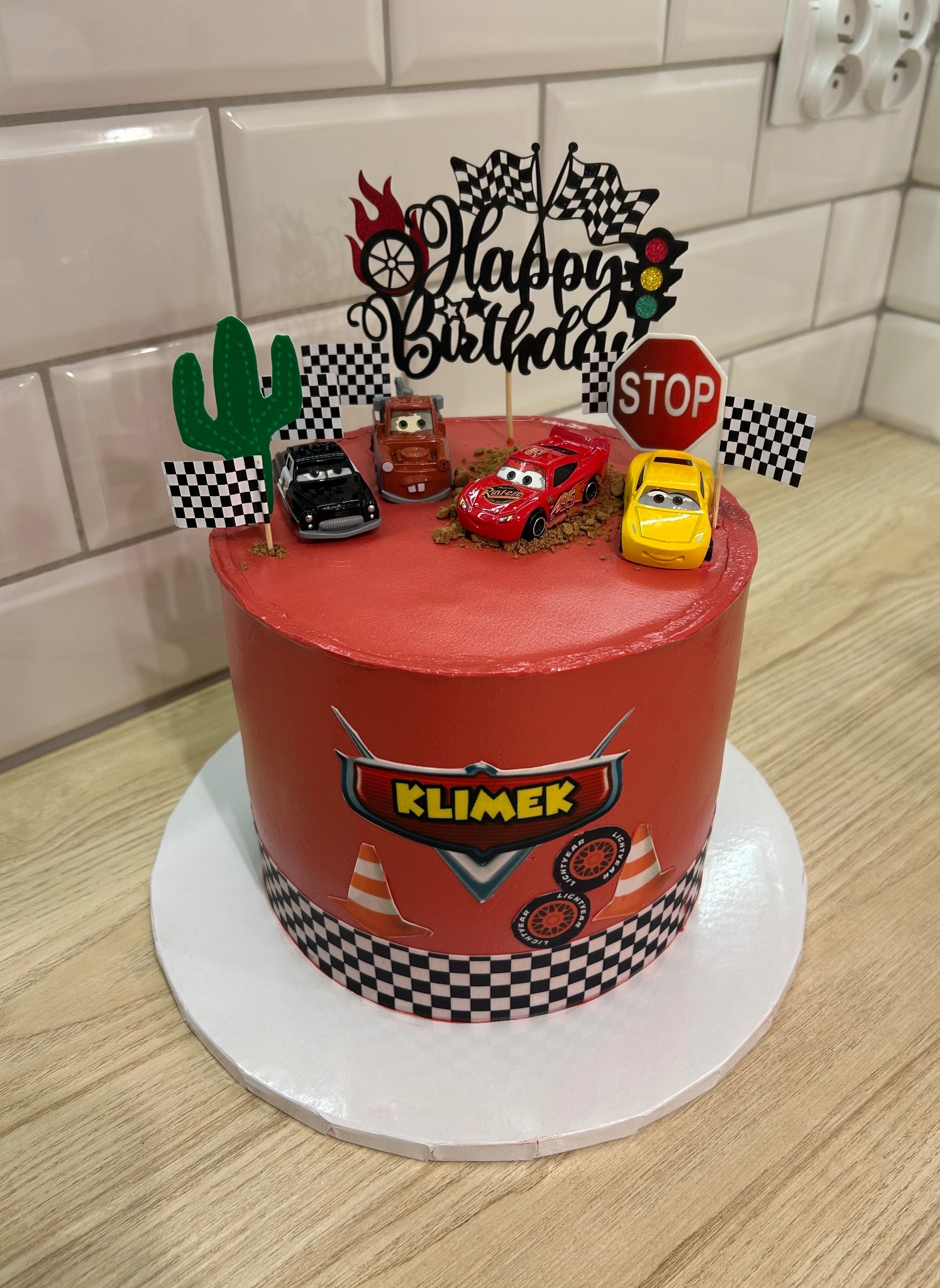 Tort urodzinowy z motywem 'Auta', czerwony krem, figurki bohaterów, dekoracje w stylu wyścigowym, na białym podeście, na drewnianym blacie.