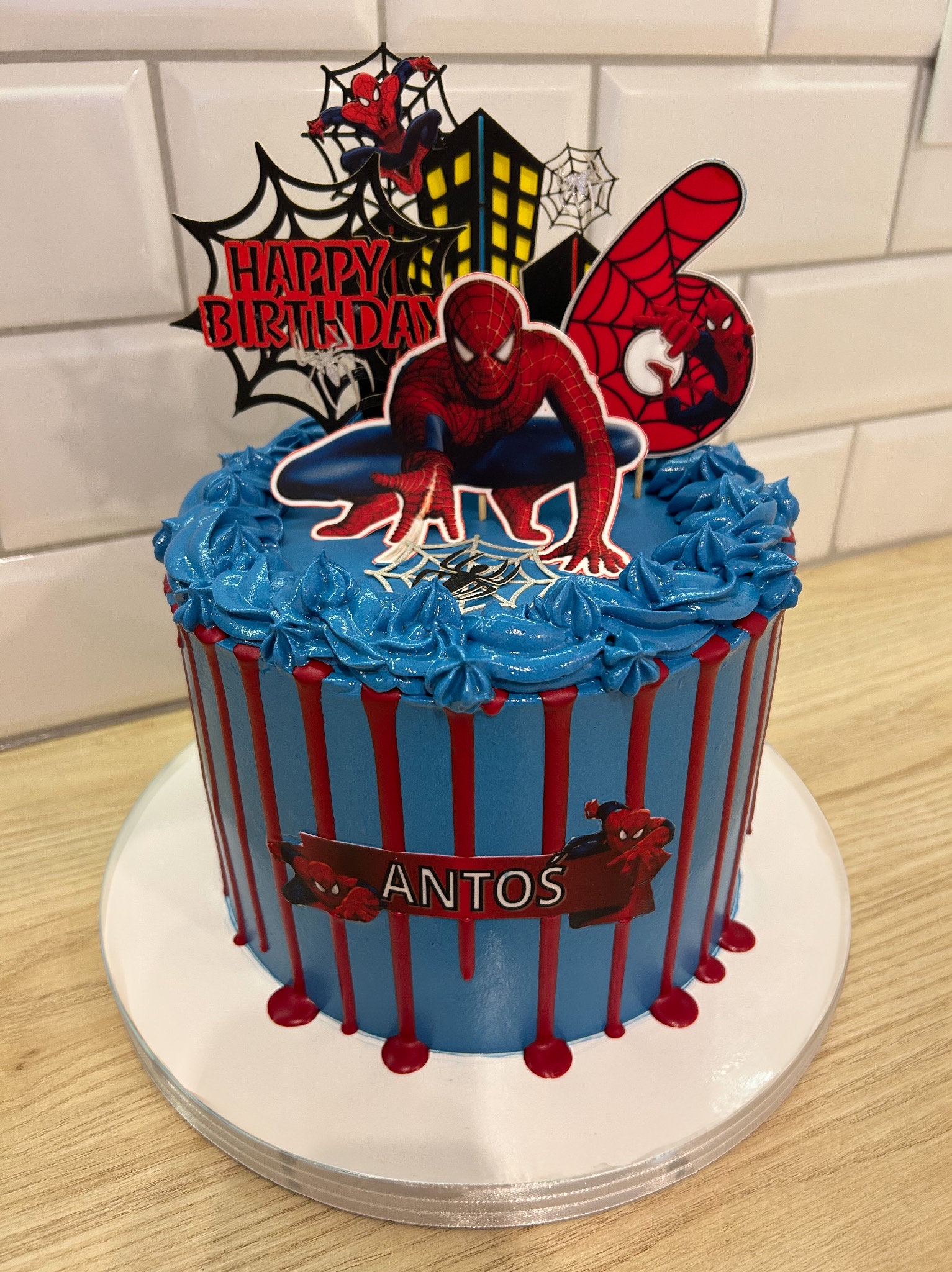 Tort urodzinowy dla Antosia z motywem Spidermana, niebieski krem, czerwone polewy, dekoracje z pajęczynami i budynkami, cyfra 6.
