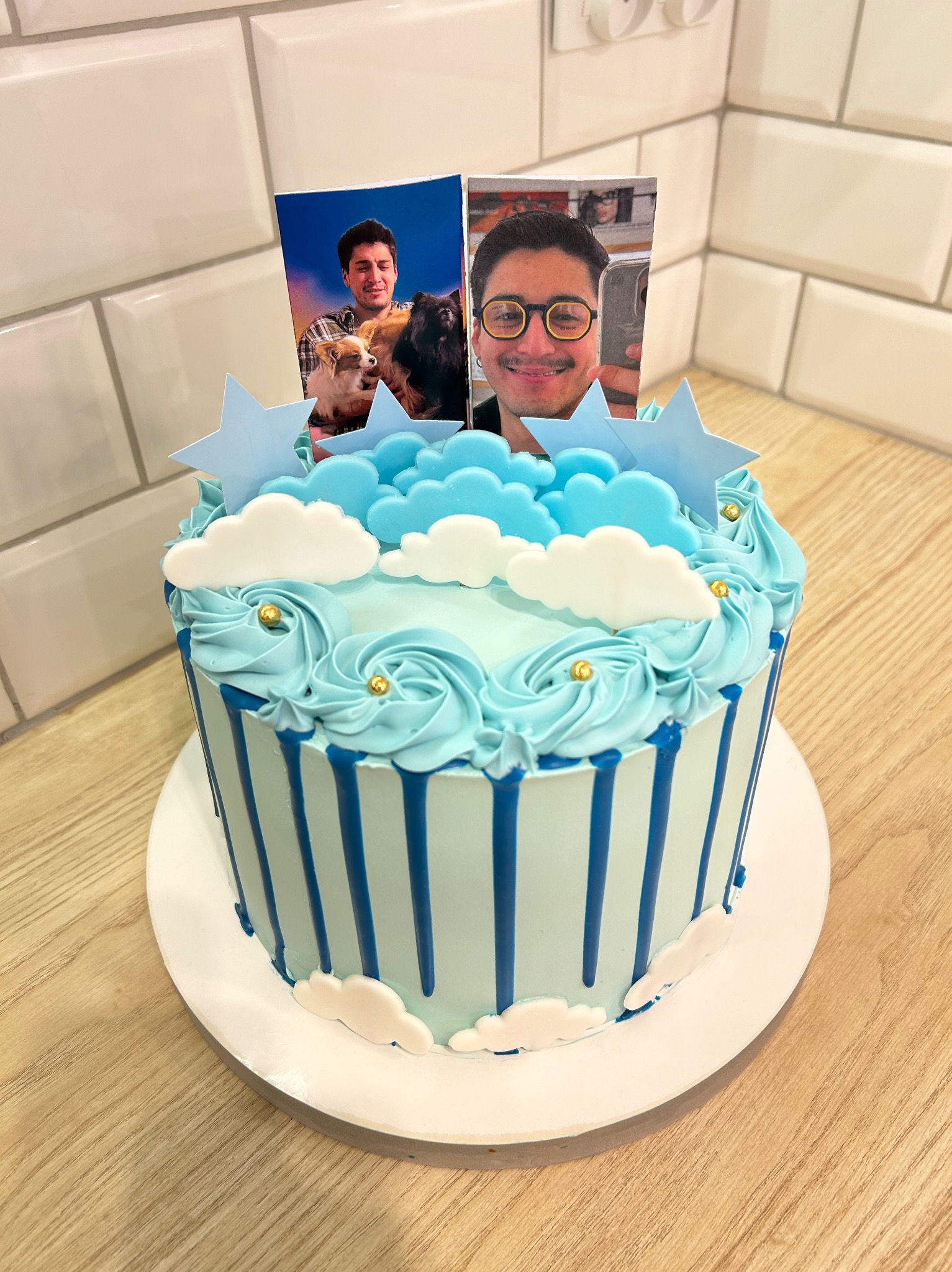 Niebieski tort z dekoracją chmur, gwiazd i zdjęciami osób, na drewnianym blacie i tle z białych płytek.