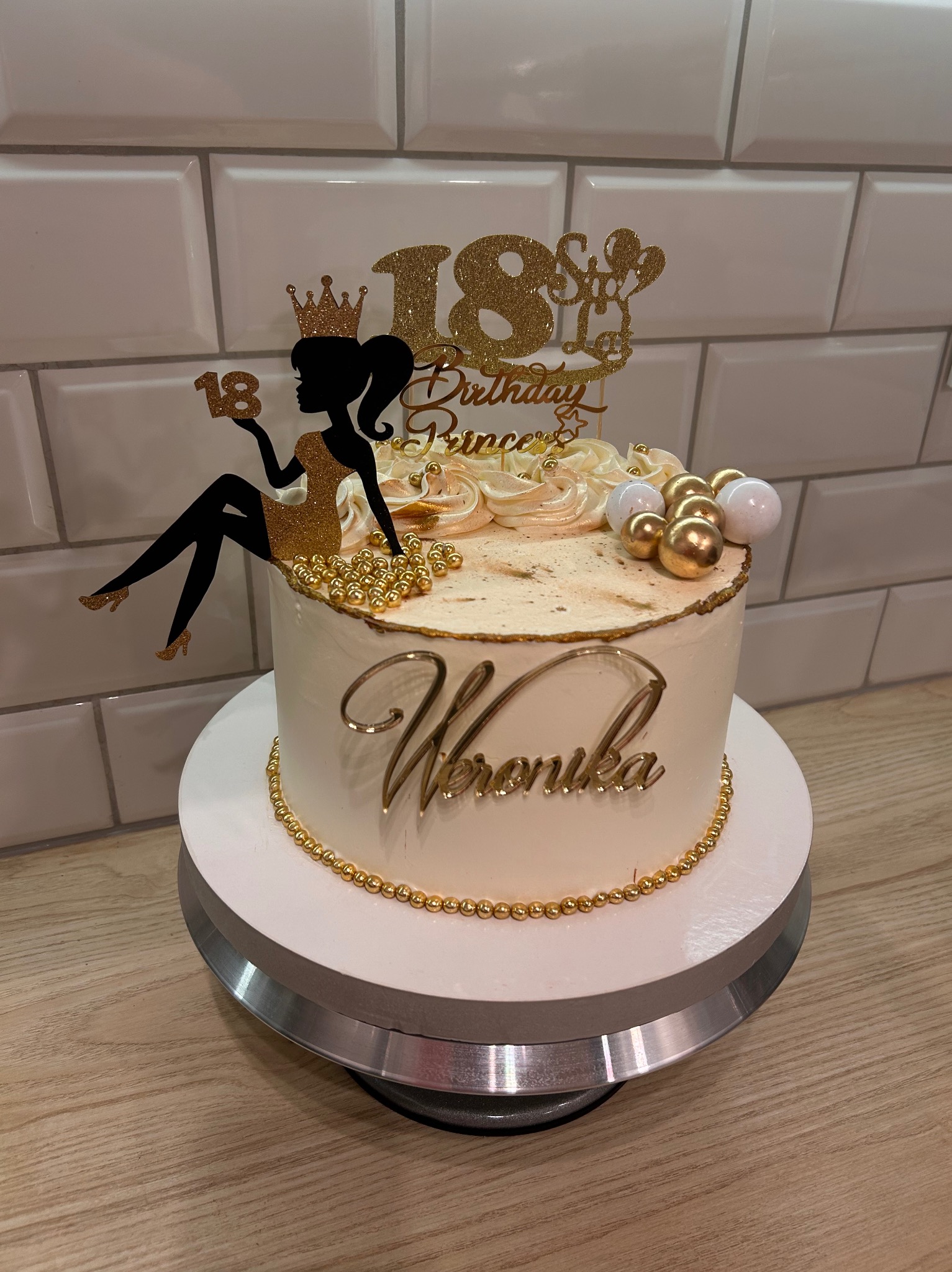 Tort urodzinowy na 18-ste urodziny z napisem 'Weronika' w złotym kolorze, dekorowany figurką dziewczyny i złotymi kuleczkami, na białym tle cegieł.