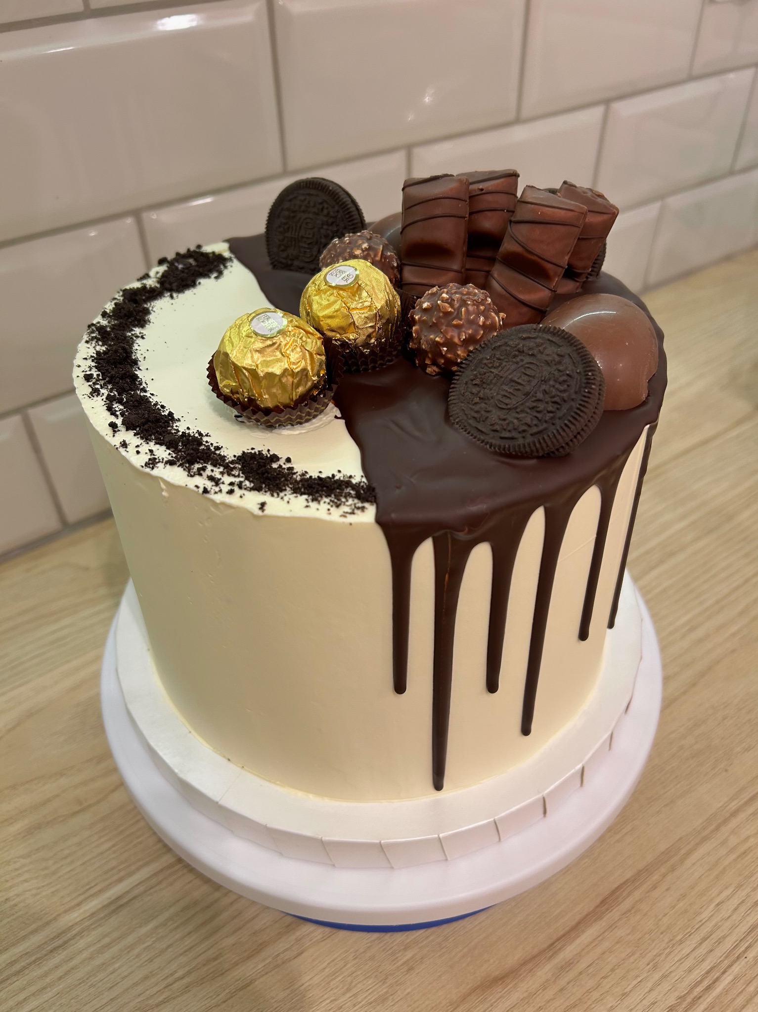 Dwukolorowy tort z polewą czekoladową, dekorowany Oreo, Ferrero Rocher i batonikami Kinder, prezentowany na białym stojaku na drewnianym blacie.