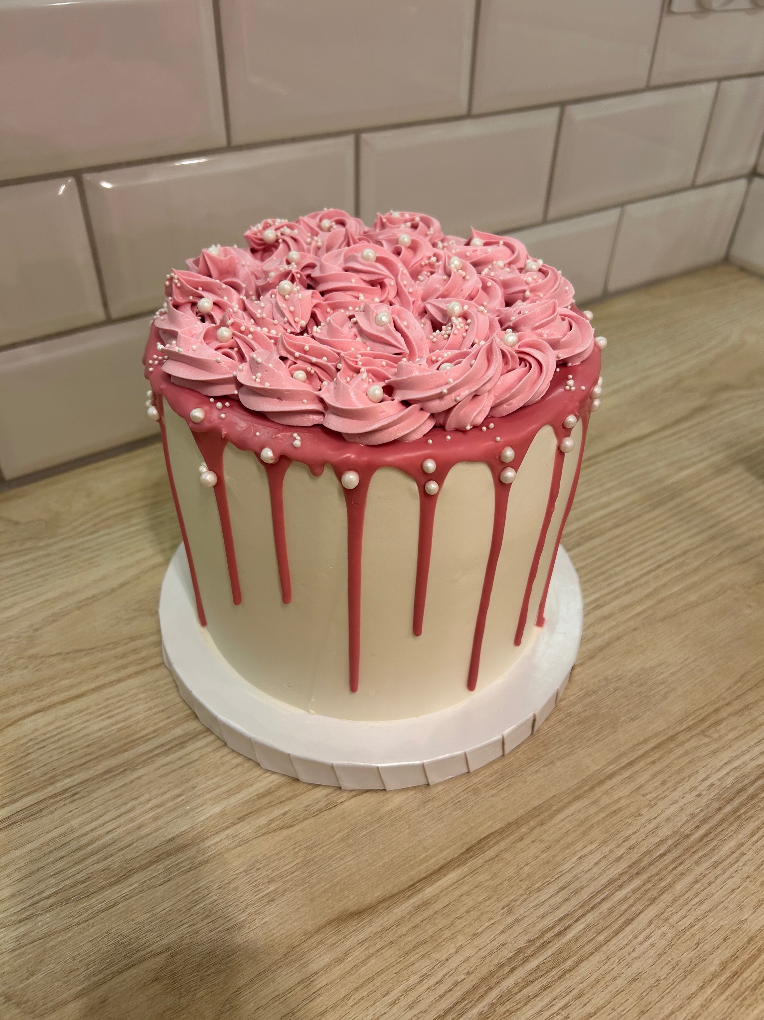 Tort z kremem różanym i perłowymi koralikami na blacie z płytek imitujących drewno, na tle ściany z białych płytek.