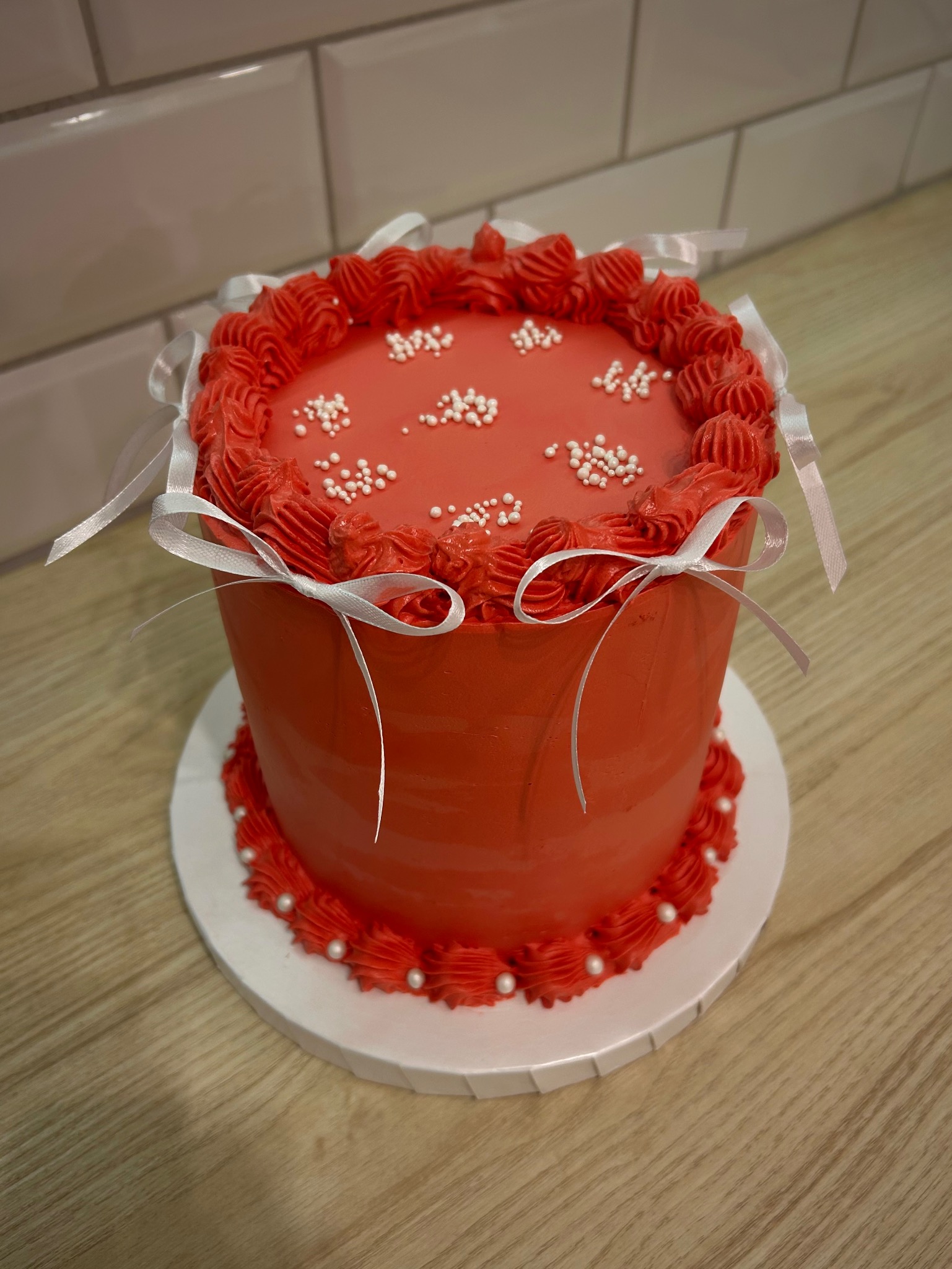 Czerwony tort z kremem i białymi perełkami, ozdobiony wstążkami, na drewnianym blacie i tle z białych płytek. Dekoracja tortu w stylu minimalistycznym.