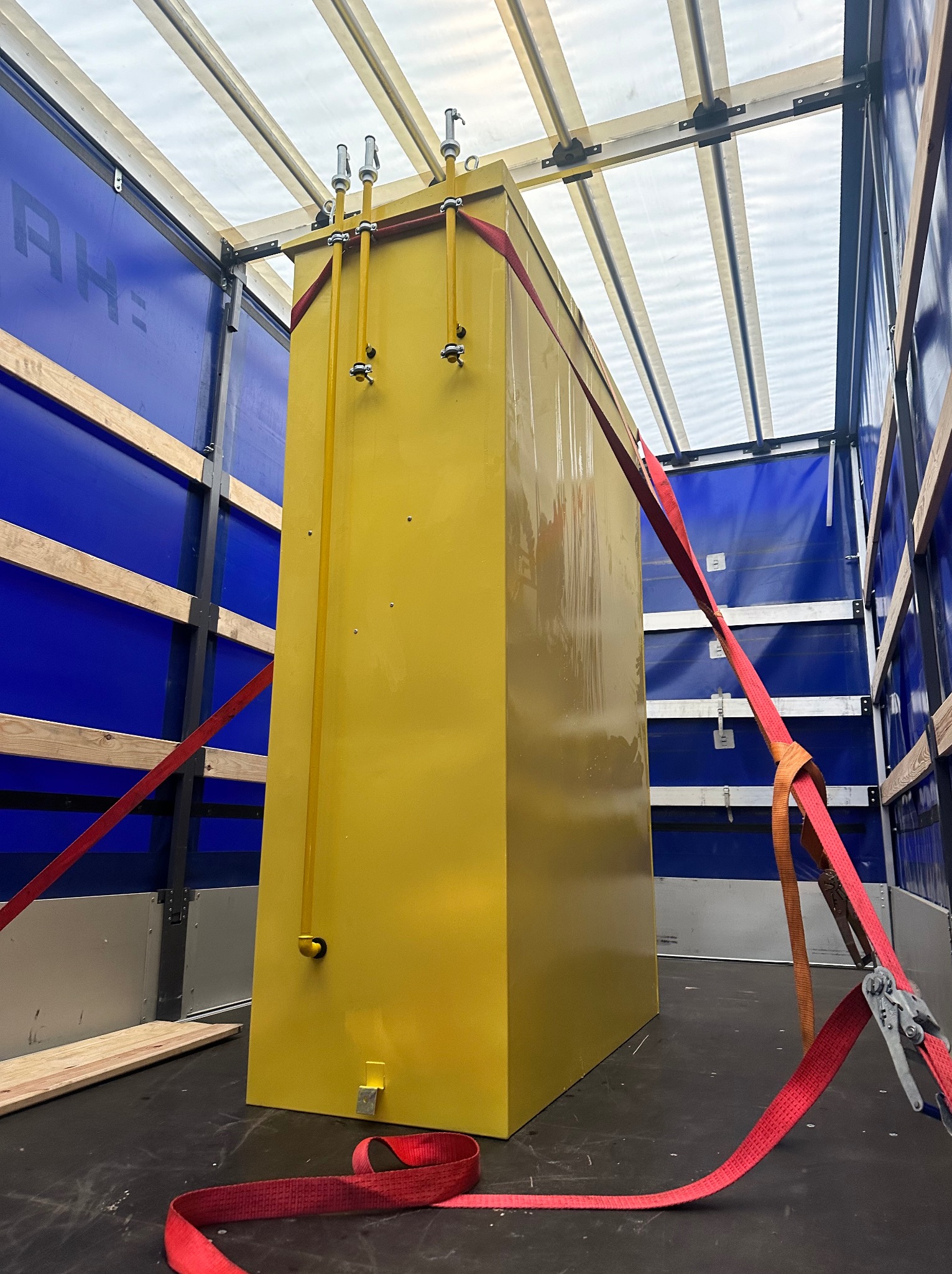 Żółty kontener zabezpieczony czerwonymi pasami w przestrzeni ładunkowej busa z niebieskimi ścianami i przezroczystym dachem, widok z góry.