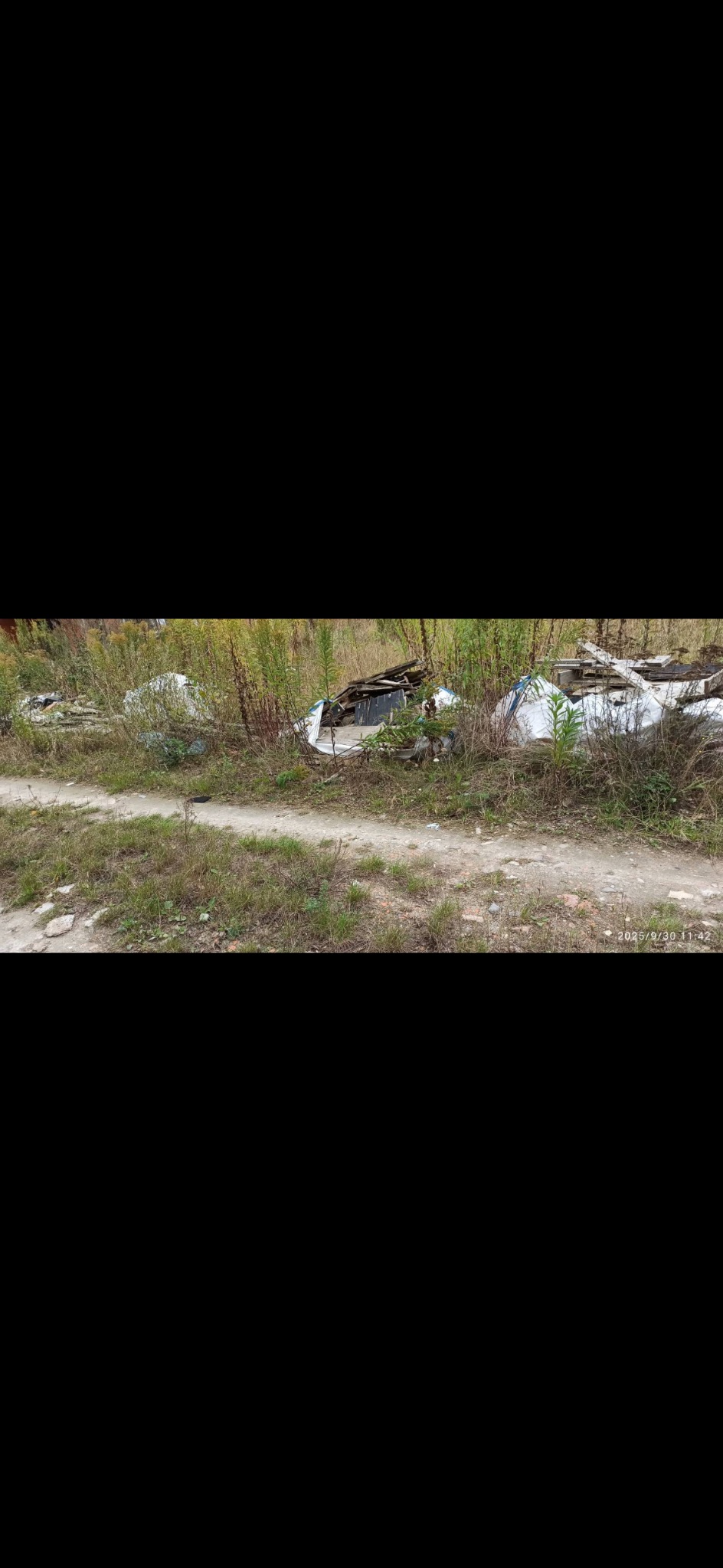 Gruzowisko na poboczu drogi, częściowo przykryte białymi plandekami, otoczone wysoką trawą i chwastami. Widoczne fragmenty drewna i innych odpadów.