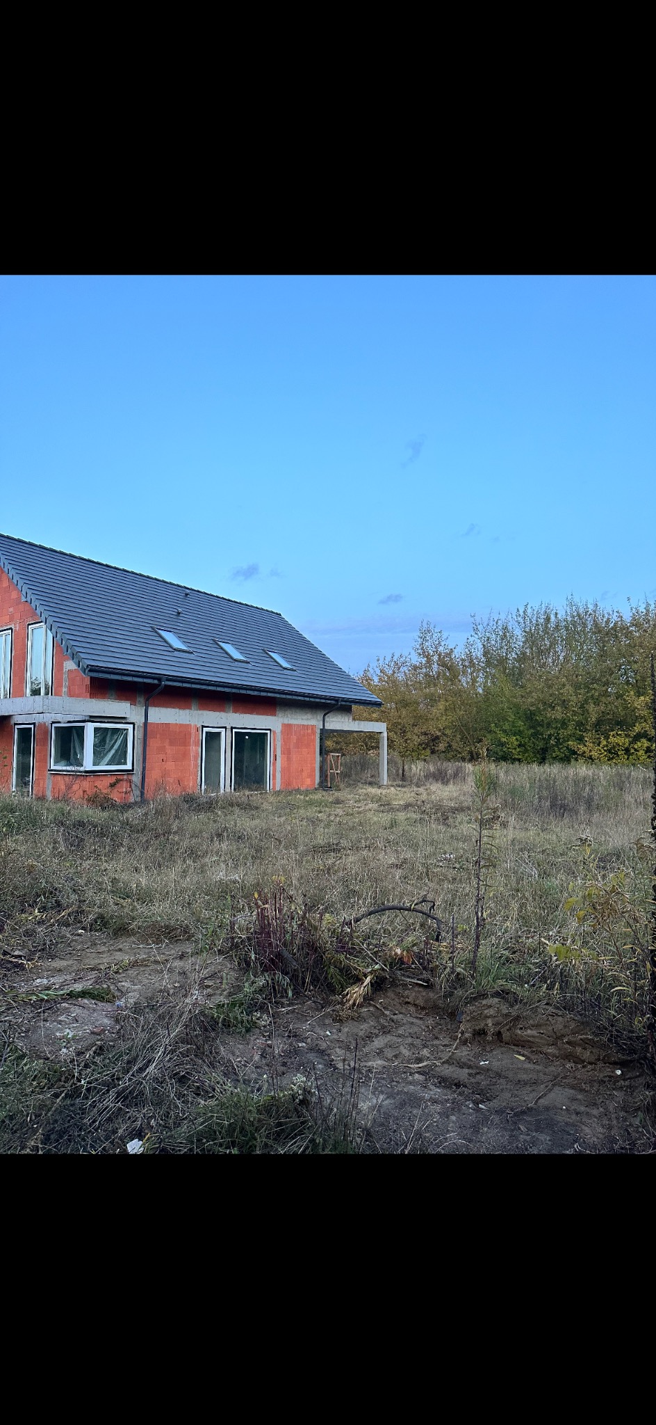 Niedokończony dom z czerwonej cegły z szarym dachem i widocznymi oknami, otoczony suchą trawą i drzewami w tle, sfotografowany w słoneczny dzień.