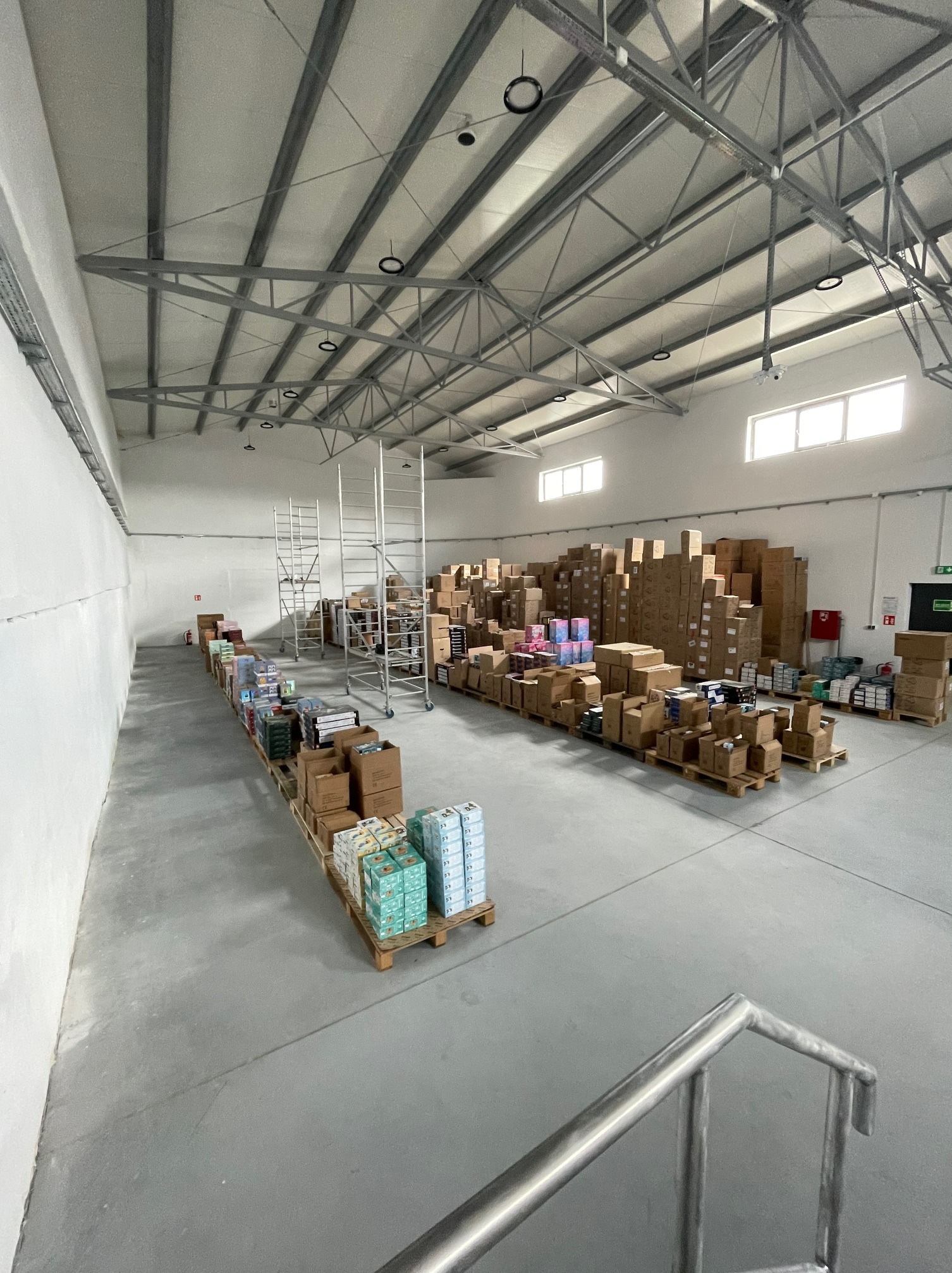 Wnętrze magazynu z paletami pełnymi kartonów i towarów, widok z góry na dużą przestrzeń z metalową konstrukcją dachu i oświetleniem.