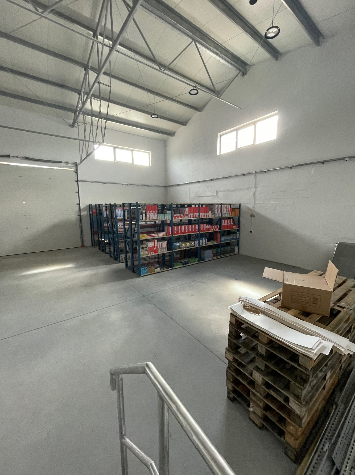 Wnętrze magazynu z regałami wypełnionymi towarem, paletami i kartonami. Hala z oknami i widoczną konstrukcją dachu.