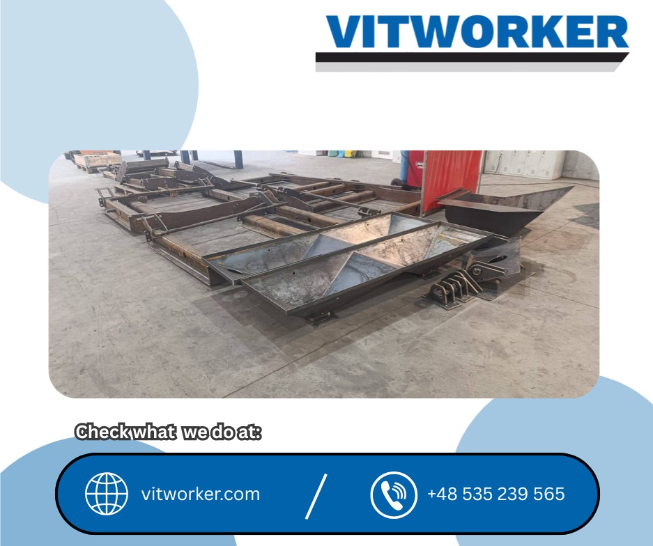 Metalowa konstrukcja hali w trakcie budowy, widoczne elementy spawane i przygotowane do montażu. Surowy beton na podłodze, w tle spawarka.