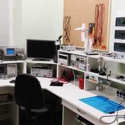 KUSZ Elektronika Krystian Kusz - Laboratorium elektroniki: stanowisko pracy z oscyloskopami, miernikami i narzędziami do naprawy urządzeń. Widoczna tablica z elementami elektronicznymi i rozdzielnica.