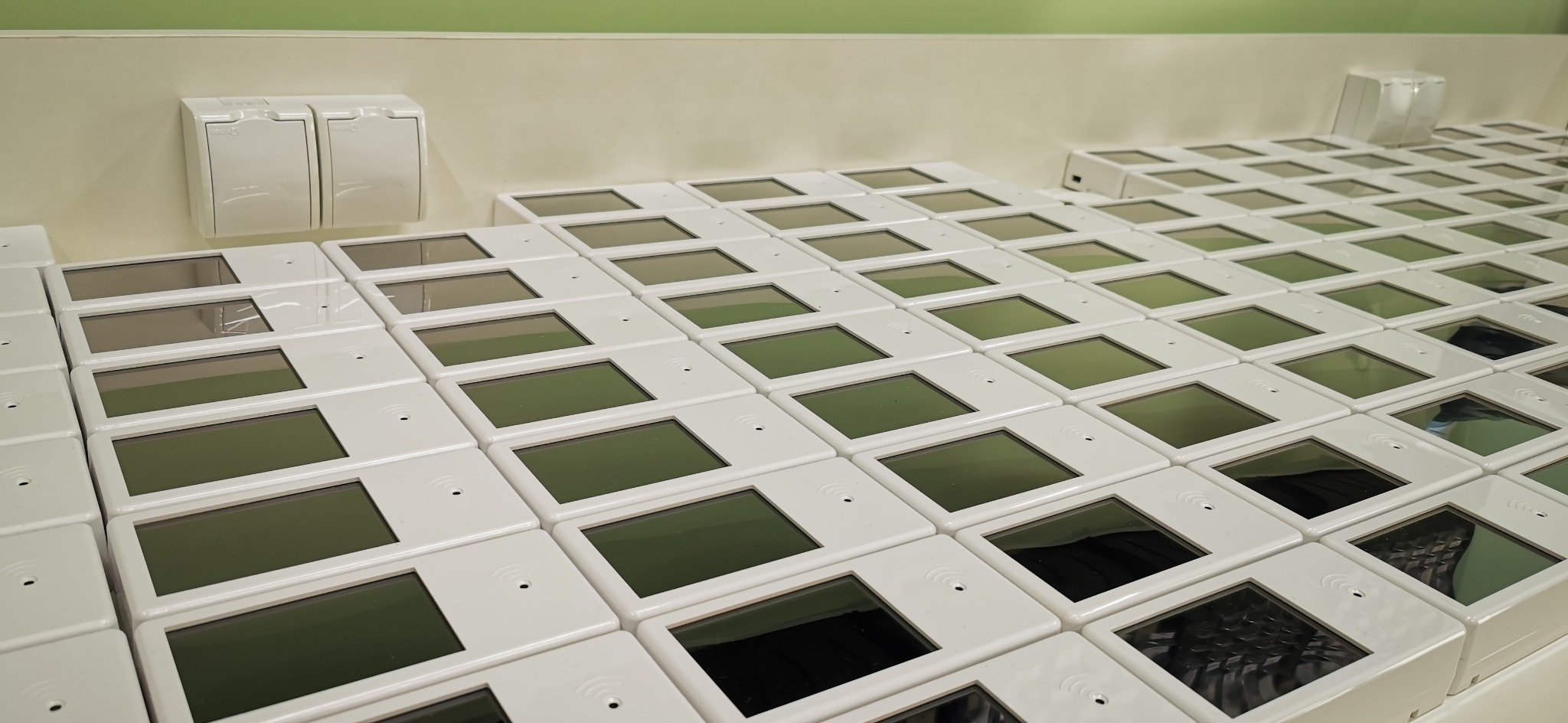 Rząd białych, kwadratowych urządzeń elektronicznych z ekranami, ułożonych na białym blacie. Widoczne gniazdka elektryczne na ścianie w tle.