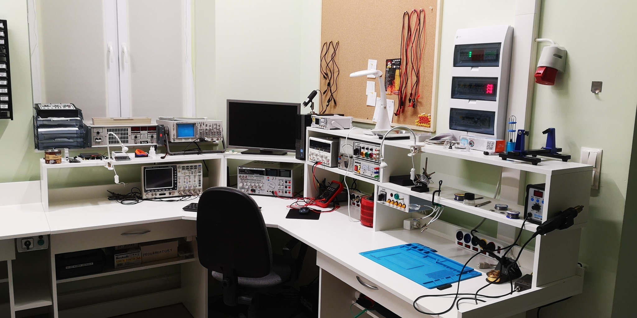 Laboratorium elektroniki: stanowisko pracy z oscyloskopami, miernikami i narzędziami do naprawy urządzeń. Widoczna tablica z elementami elektronicznymi i rozdzielnica.