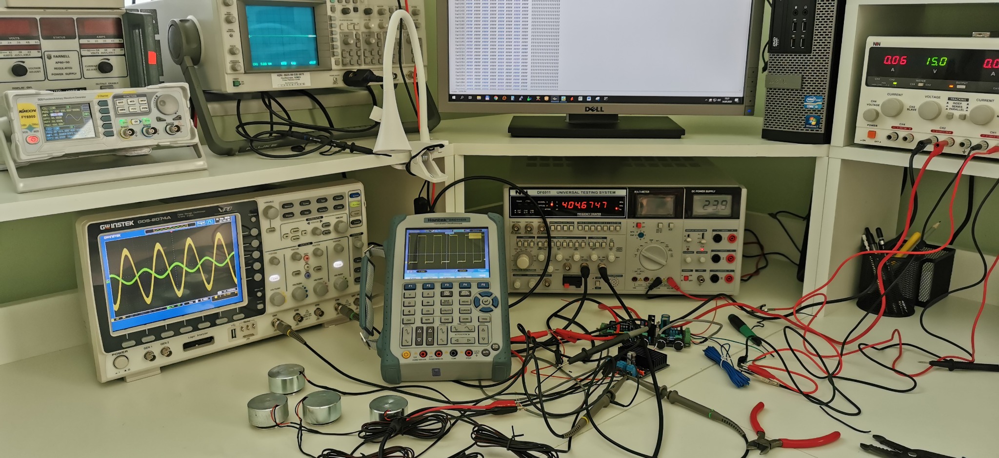 Stanowisko pracy elektronika: oscyloskopy, zasilacz, multimetr, prototyp układu scalonego połączony przewodami, monitor z kodem. Uruchamianie i testowanie prototypów.
