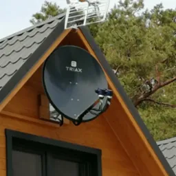 Antena satelitarna Triax i antena naziemna na dachu drewnianego domu z oknem. Widok z dołu na tle nieba i drzew.