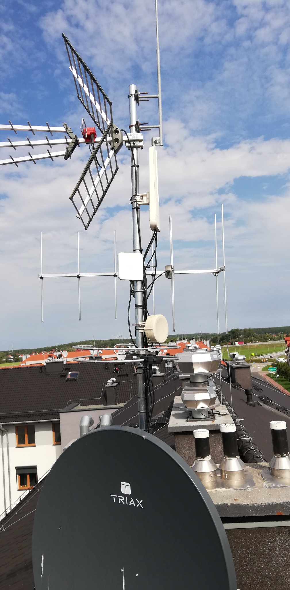 Anteny różnego typu, w tym satelitarna Triax, zamontowane na dachu budynku z kominami na tle błękitnego nieba z chmurami i zabudowań.