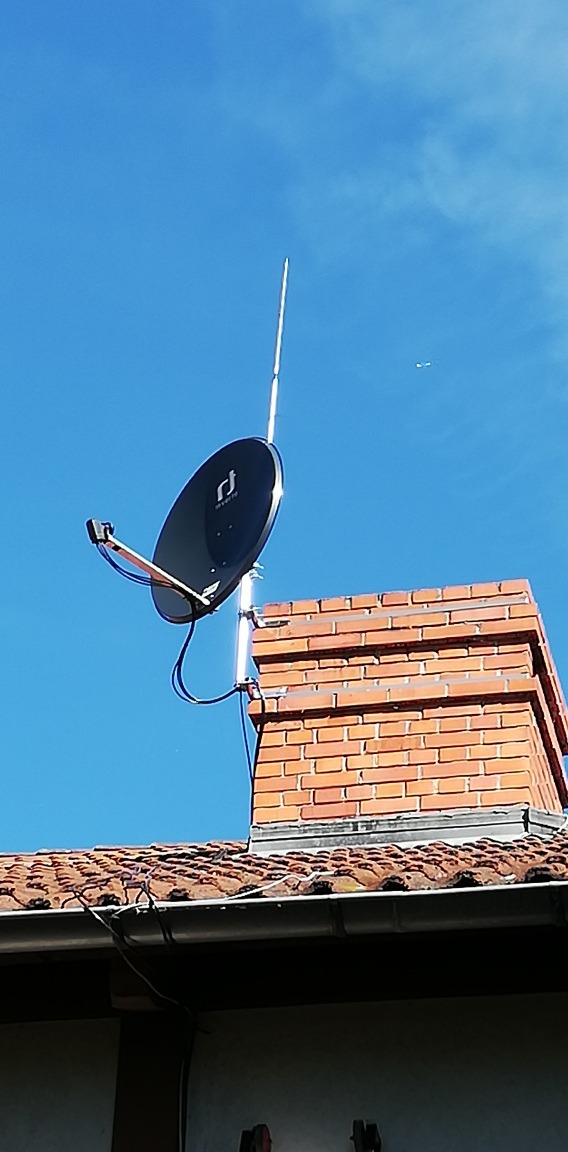 Antena satelitarna z długim masztem zamontowana na kominie z czerwonej cegły, na tle błękitnego nieba. Widoczny fragment dachu z dachówką.