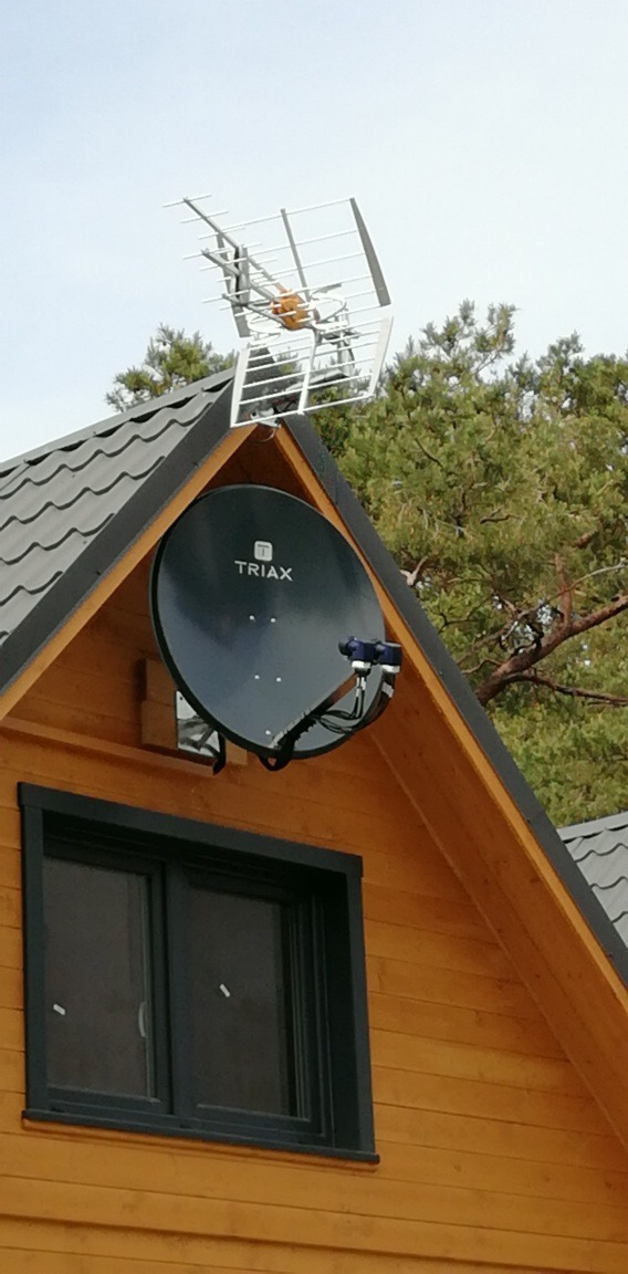 Antena satelitarna Triax i antena naziemna na dachu drewnianego domu z oknem. Widok z dołu na tle nieba i drzew.