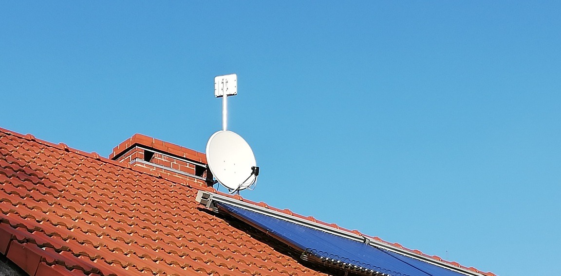 Antena satelitarna i antena LTE zamontowane na dachu z czerwoną dachówką i panelami solarnymi, na tle błękitnego nieba.