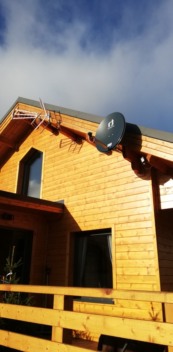 Zewnętrzny montaż anteny satelitarnej i anteny naziemnej na drewnianym domu jednorodzinnym. Widok z dołu na tle błękitnego nieba.
