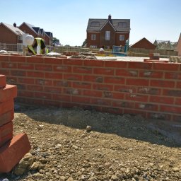 OPTIROOMS SERVICE & CONSTRUCTION SPÓŁKA Z OGRANICZONĄ ODPOWIEDZIALNOŚCIĄ - Ceglana ściana w trakcie murowania, z widocznym pracownikiem w kasku i stosem cegieł. W tle nowe domy w budowie.